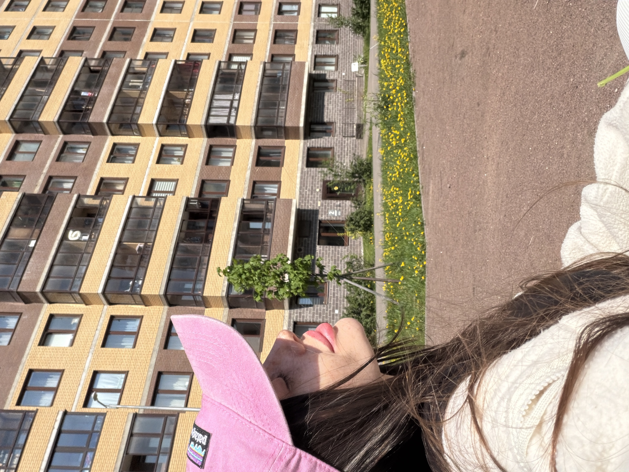 The image shows a woman wearing a pink baseball cap and a white hooded sweatshirt, lying on a brown surface next to a patch of yellow flowers. The background reveals a large apartment building with brown and yellow brick facades and multiple rows of windows. The location is Saint Petersburg, Russia, as evidenced by the architectural style of the building.