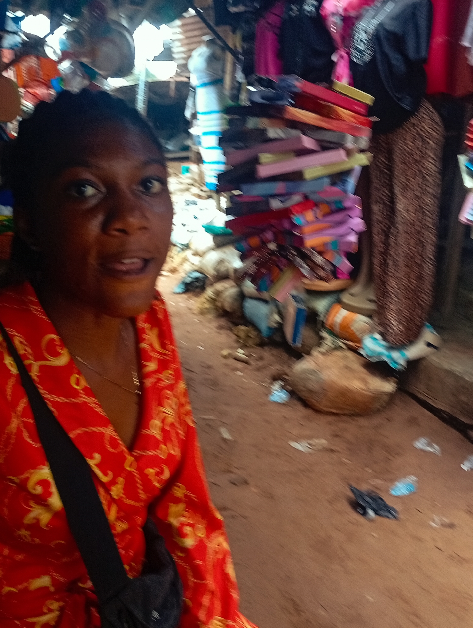 This is a close-up, eye-level, medium shot of a young Black woman taken in an outdoor market in Nkpor, Nigeria. The woman is in the foreground, facing the camera with her mouth slightly open. She is wearing a bright red top with a gold ornate pattern. A black strap, likely from a bag, crosses her chest. The background is blurred, showing stalls with various goods, including stacks of colorful fabric or clothing and what appear to be coconuts or other produce on the ground. The ground is reddish-brown dirt. The scene appears to be during the day, with bright ambient light. The woman's expression is difficult to discern due to the blur and the angle, but she seems to be looking directly at the camera. There is no visible text in the image.
