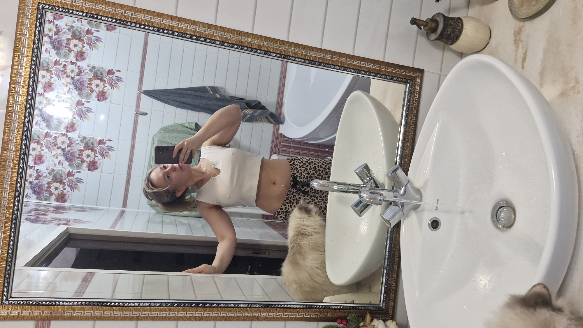 The image shows a bathroom scene reflected in a mirror with a patterned gold frame. A woman is reflected in the mirror, holding a smartphone and appearing to take a selfie. She is wearing a cropped white top and leopard-print bottoms. Behind her, in the reflection, are patterned floral tiles and white wall tiles, a bathtub, and towels. In the foreground is a white sink with running water. Part of a fluffy cat is visible near the bottom of the sink. The bathroom appears to be well-lit. There is no clear indication of the location being Otchor, Russia, or any other specific city.
