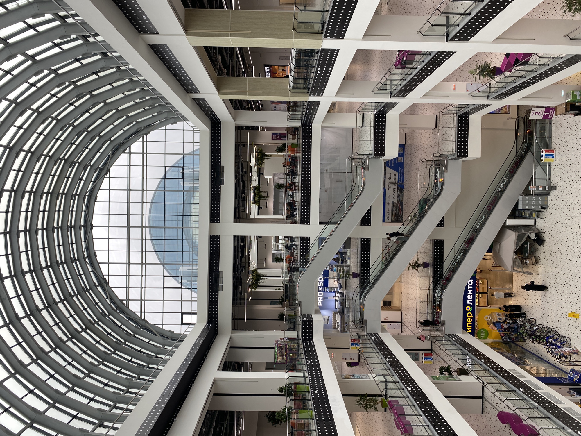 The image depicts the interior of a modern, multi-level shopping mall with a large, arched glass ceiling. The mall features multiple floors with escalators connecting them. Balconies with black decorative borders and white circular perforations line the sides of each level, offering views into the central atrium.
On the ground floor, there are several shops, including one with a large yellow sign that reads "Империя" (Imperiya) and another that says " FRESH COFFEE". Bicycles are parked in a row near the entrance of the "Империя" store, and a temporary tent structure is set up nearby. Several people are visible walking around on different levels, and some are seated at tables in a food court area on an upper level. Plants are used as decorative elements throughout the mall.
The architectural style is contemporary, with white pillars, minimalist design, and extensive use of glass and metal. The overall impression is one of a bright, spacious, and well-lit commercial environment.
The location is identified as the "Novokuybyshevsk City Mall" in Novokuybyshevsk, Russia.