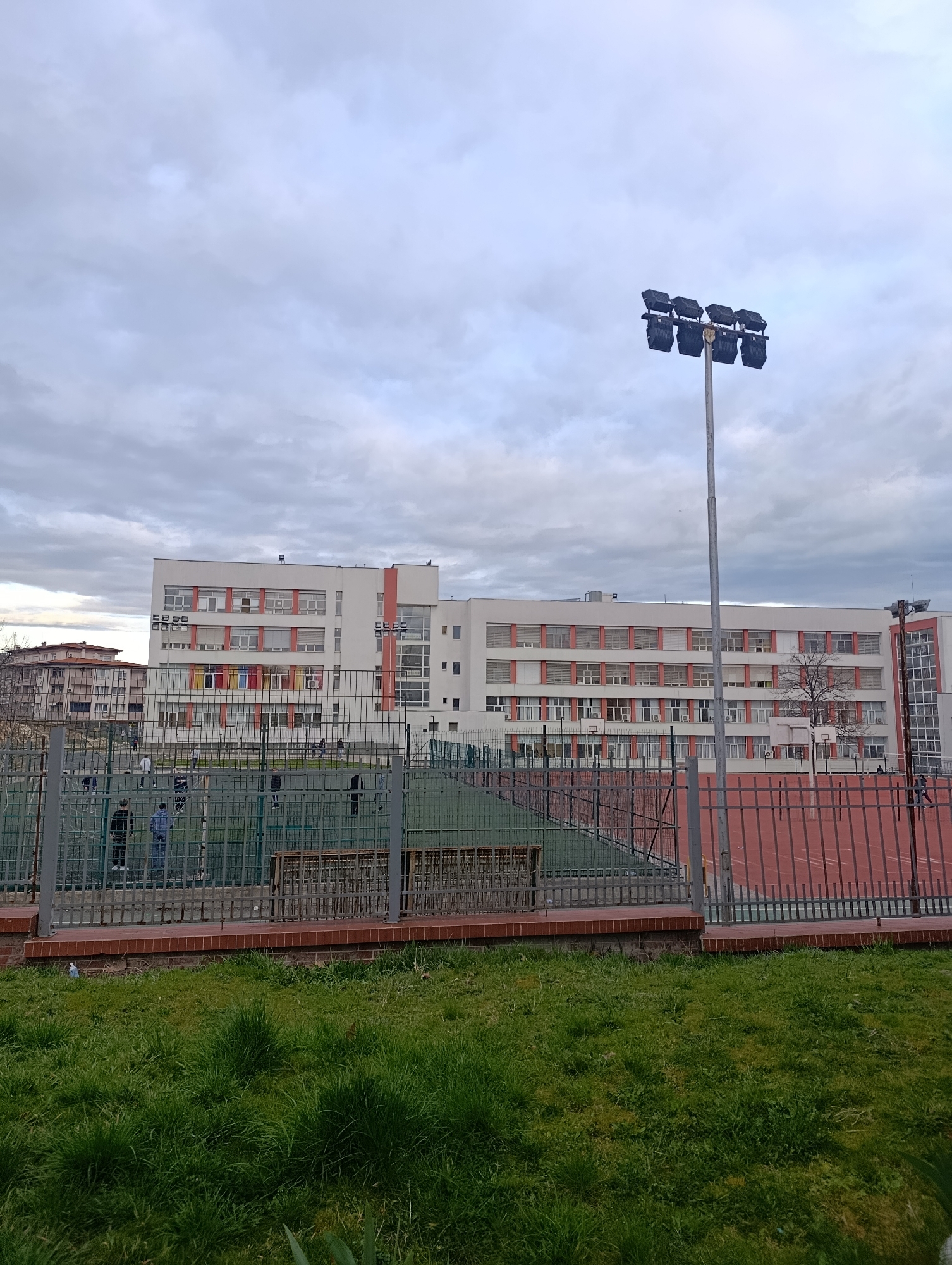 The image shows a multi-story building with a white facade and coral-colored accents on window frames and vertical stripes. The building is located in Kardzhali, Bulgaria. A sports field or playground is situated in front of the building, enclosed by a gray metal fence. Within the field, there appear to be people, possibly engaged in sports activities. A tall light pole with multiple spotlights is positioned to the right of the frame. A patch of green grass fills the foreground. The sky is overcast with gray clouds.
