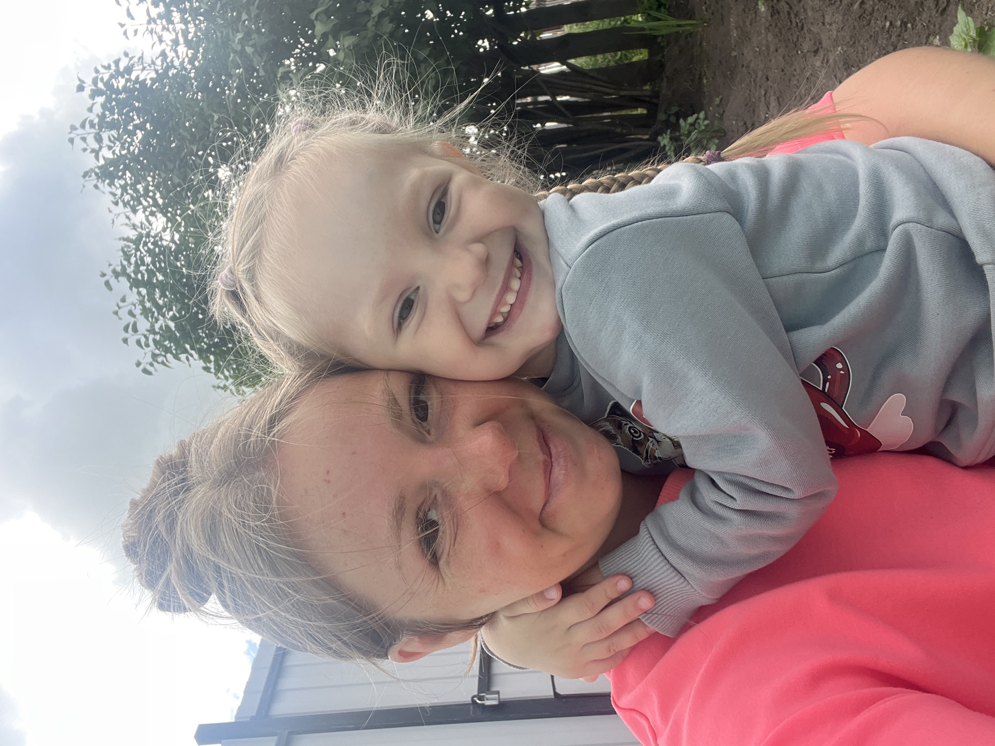 A close-up selfie of a woman and a young girl embraces outdoors. The woman, with her hair tied up in a bun, wears a bright pink sweatshirt. The girl, with blonde hair in braids, is wearing a light blue sweatshirt and is hugging the woman. They are both smiling. In the background, there is a fence, trees, and a cloudy sky. The image appears to be taken in a residential area, possibly a backyard. The location is identified as Sukhinskoe, Russia, suggesting a rural or suburban setting.