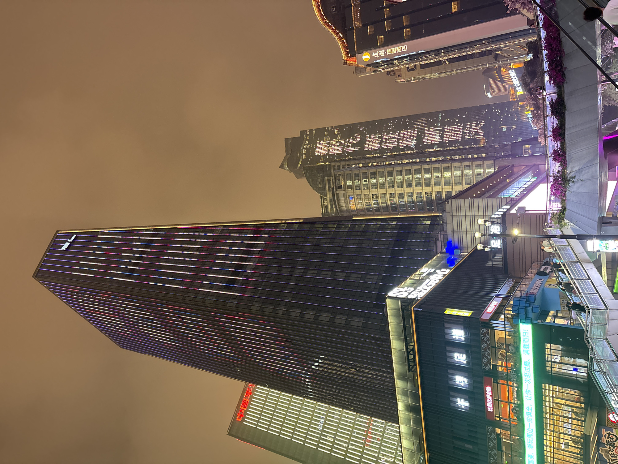 The image presents a nighttime scene in the Yuzhong District of China, featuring several high-rise buildings illuminated against a hazy sky. The dominant structure is a tall skyscraper adorned with horizontal lines of white, red, and blue lights, stretching towards the top left corner of the frame. Adjacent to this building is another high-rise with visible windows and illuminated signage, including Chinese characters.
In the foreground, there's a building with multiple levels, likely a commercial space, with glass windows revealing interior scenes and various advertisements or signage. The OCR text indicates the presence of businesses like "Big Pizza Buffet" and a cultural center.
The environment suggests an urban setting, with the light and sky indicating some form of air pollution or fog, common in large cities. A pedestrian walkway or commercial area is subtly implied in the immediate foreground.