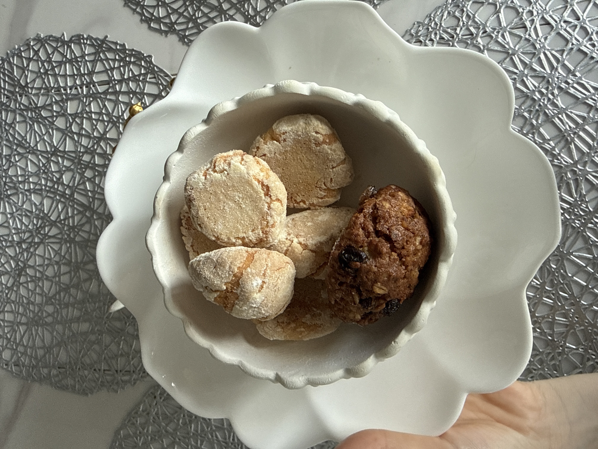 A high-angle, close-up shot shows a white decorative bowl filled with several cookies. The bowl sits on a larger white decorative dish, also in the shape of a flower. On the left and right of the dishes, portions of a silver woven-material placemat are visible. The cookies appear to be of two varieties: light-colored, dusted with powdered sugar; and a dark, raisin-studded cookie. A person's hand is partially visible at the bottom edge of the image, supporting the bowl. The location is not discernible from the image content, and there is no information to connect the image to Khimki, Russia.