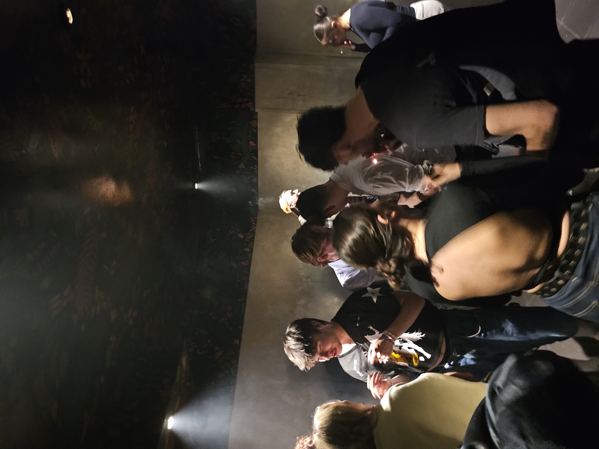 A group of people are gathered closely together, seemingly looking at something on the ground or at a low level. They are in a dark, enclosed space, possibly indoors. There is concrete and lighting in the space. The location is Paris, France.