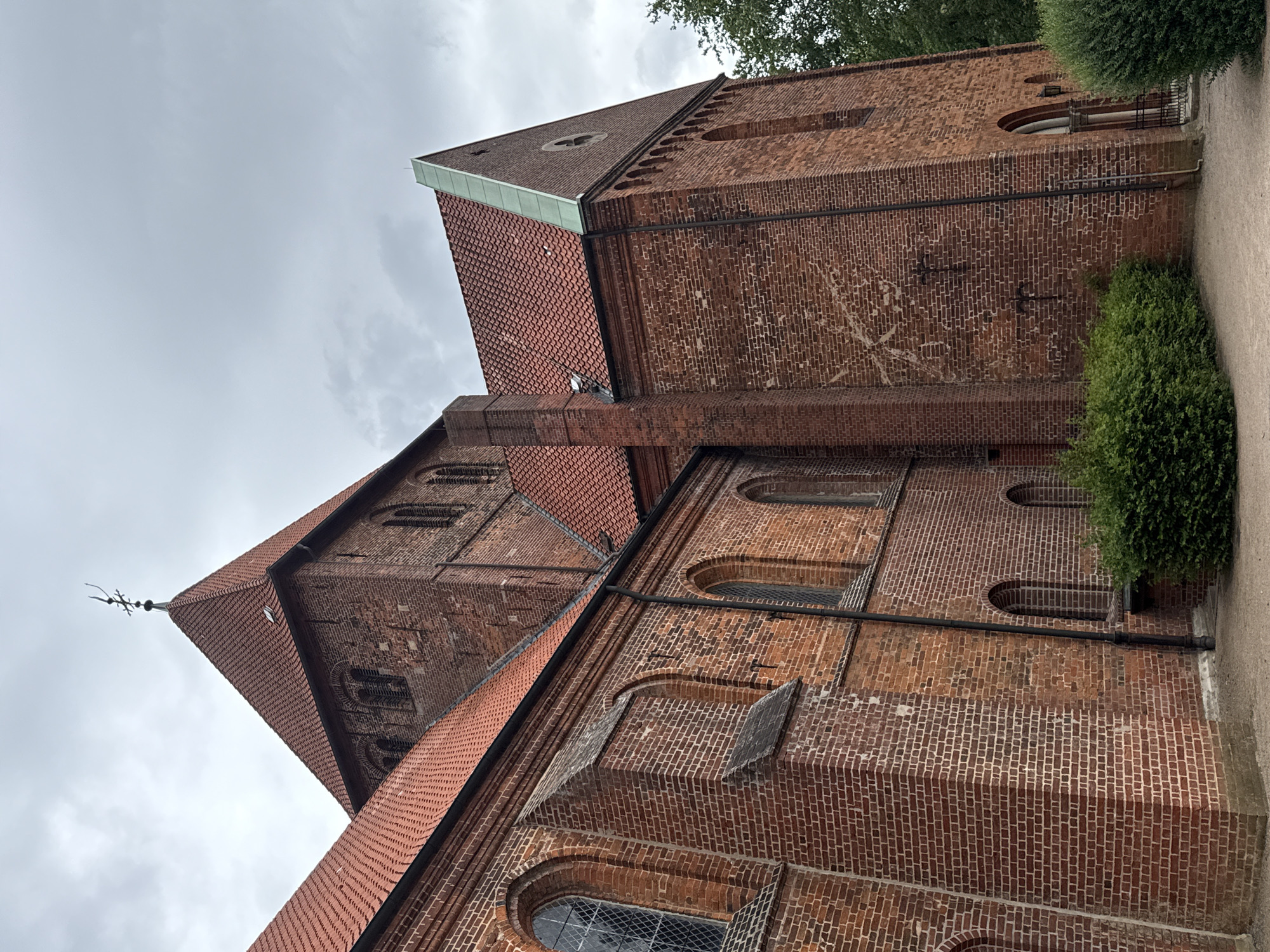 The image shows the exterior of the St. Marien Church in Bassum, Germany. The church is constructed of red brick, with a steeply pitched red tile roof. Several arched windows are visible on the brick facade. A tall, square tower with a pointed roof topped by a weather vane rises from the center of the church. Lush green bushes are planted in front of the building, and a gravel path leads to an entrance. The sky above is overcast.