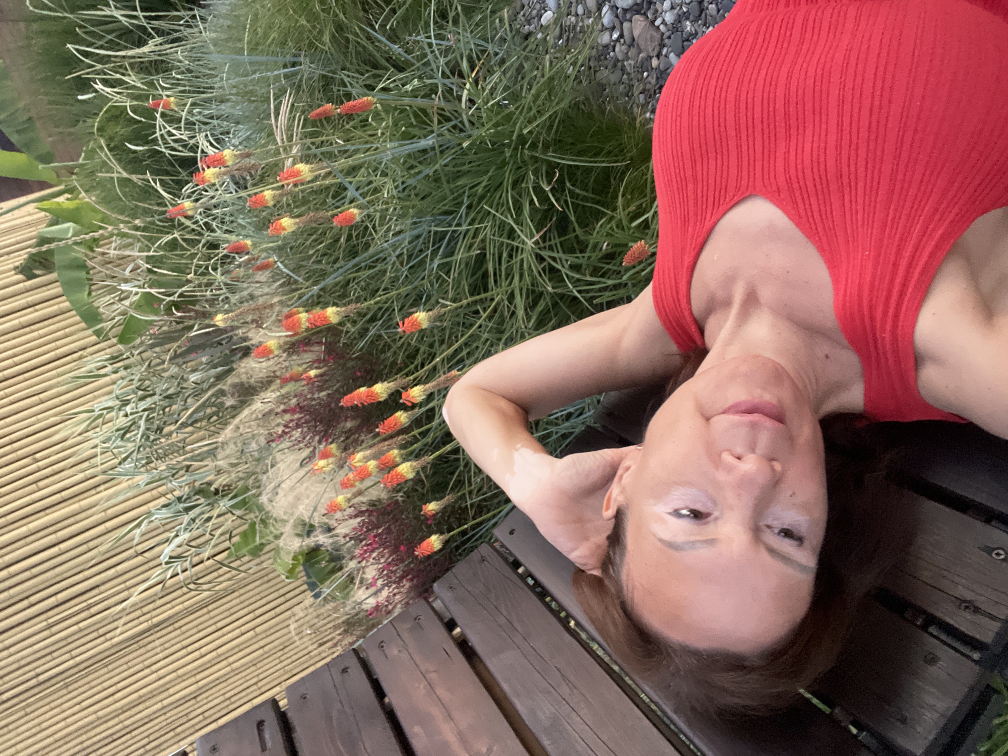 The image shows a woman lying on a wooden bench in an outdoor setting, possibly a garden or park, with a bamboo fence in the background and lush green foliage with red and yellow flowers. The woman is wearing a red ribbed top and her left arm is raised behind her head. She has brown hair and appears to be in her late 40s or early 50s. The image is taken from a high-angle perspective, looking down at the woman. The environment suggests a warm, possibly tropical climate, consistent with locations like Sochi, Russia, known for its botanical gardens and resort areas. The overall impression is a relaxed and intimate portrait.