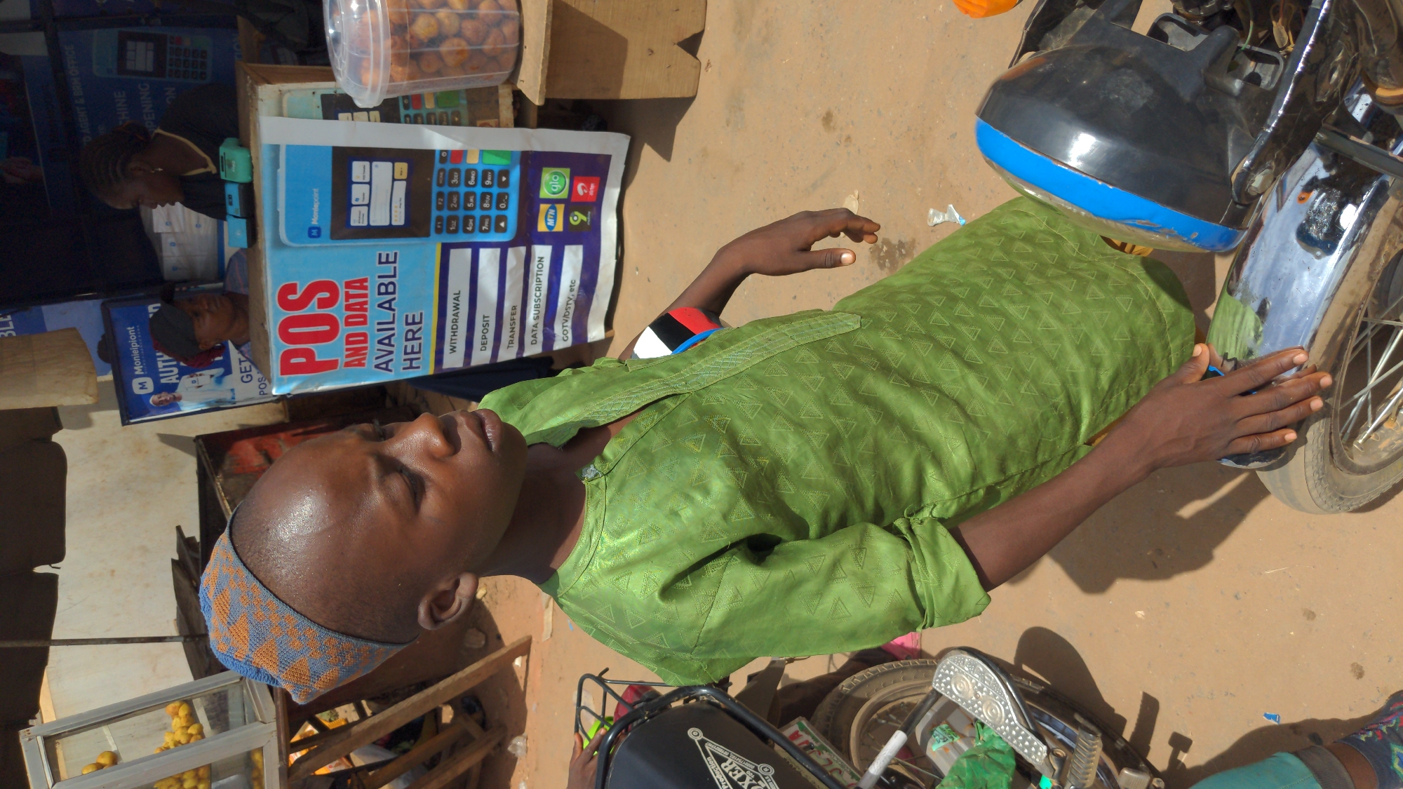 The image shows an individual in Ado, Nigeria, wearing a green patterned garment, a blue and orange patterned cap, and a red, white, and black bracelet. They are positioned adjacent to a motorbike and a POS machine advertisement, which indicates money and data services are available. Also, there is a display of food items. The scene takes place outdoors on a dirt surface.