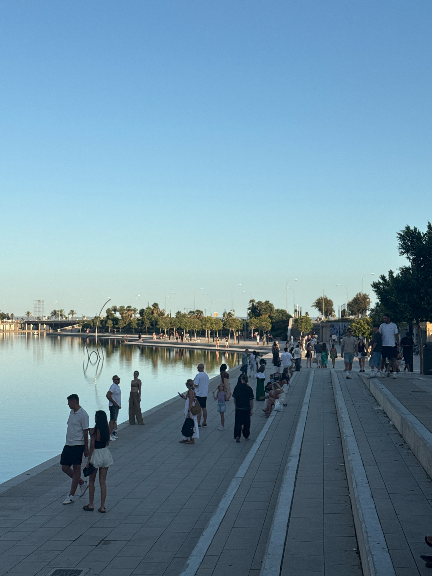 The image depicts a waterfront promenade in Palma, Spain, likely the Parc de la Mar, under a clear blue sky. A calm body of water borders the left side of the frame, reflecting the sky and surrounding trees. On the paved promenade, a diverse group of people are engaged in various activities: walking, standing, and sitting. Several individuals are walking along the path, some in pairs, others alone. Others are gathered in small groups, conversing or observing the scenery. In the background, lush greenery with palm trees lines the opposite side of the water, and modern architectural structures, including lampposts and a bridge, are visible. The overall atmosphere suggests a leisurely day out in a public urban space.