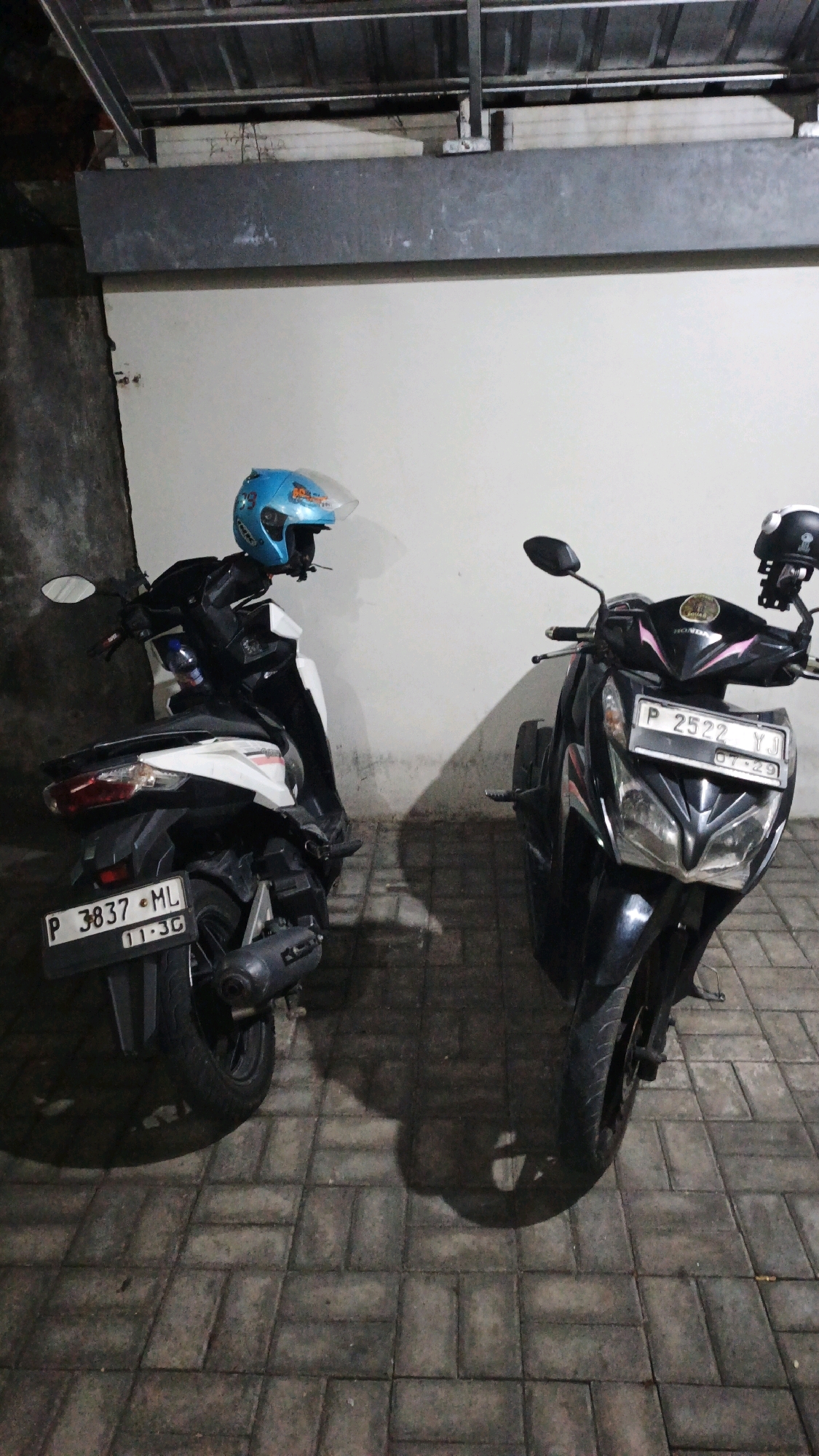 Two parked motorcycles are visible in the image, set against a light-colored wall. The motorcycle on the left is white and black, with a blue helmet resting on its handlebars. Its license plate reads "P 3837 ML 11-3G". The motorcycle on the right is black, with a black helmet on its handlebars. Its license plate reads "P 2522 YJ 07-29". The ground is paved with grey rectangular tiles. The scene appears to be in an outdoor, covered area, possibly a parking space in Denpasar, Indonesia, given the license plate format.