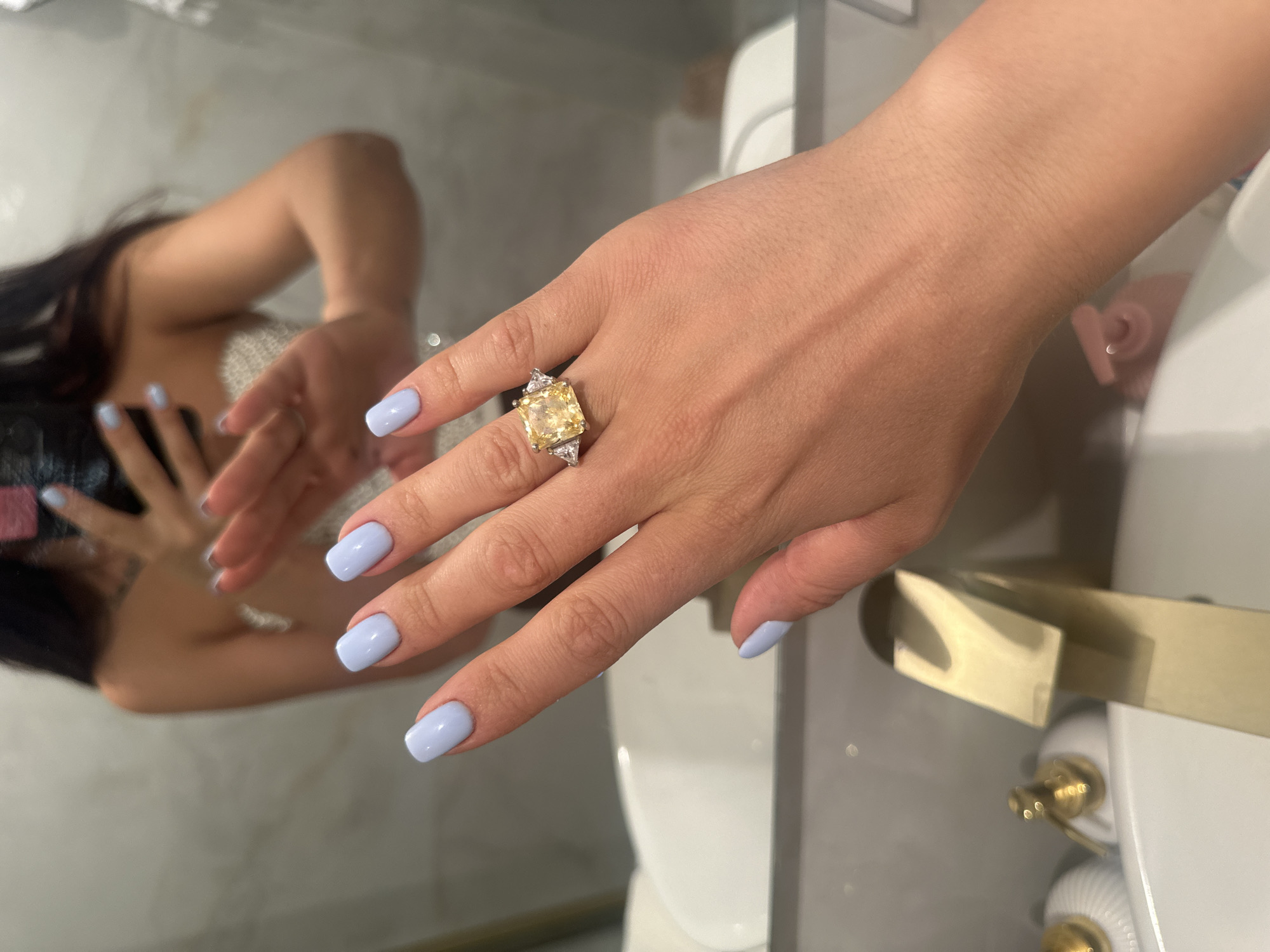 A light-skinned woman's right hand, adorned with a large yellow square-cut gemstone ring flanked by smaller triangular stones, is prominently displayed. The woman's nails are painted a pastel blue color. Her reflection is visible in a nearby mirror, showing her holding a phone, suggesting she is taking a picture. The background reveals a bathroom setting, with a white toilet and gold-toned hardware visible. There is no clear indication that this photo was taken in Khimki, Russia.