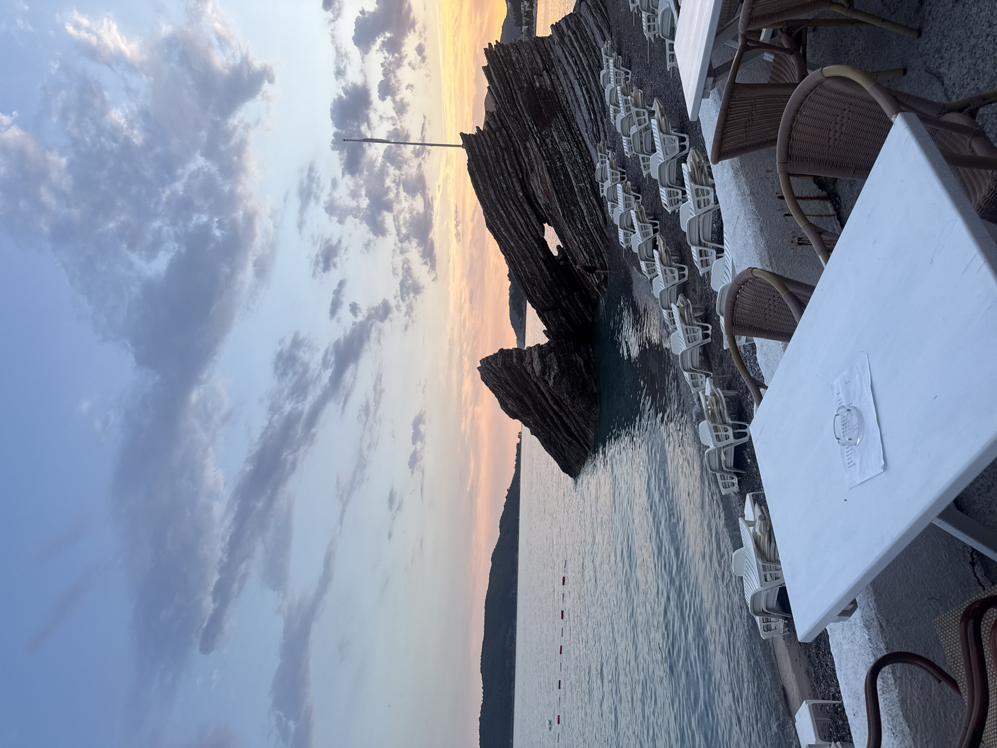 The image depicts a serene beach scene at dusk in Bečići, Montenegro. The foreground features a white table with wicker chairs, suggesting an outdoor seating area for a cafe or restaurant. Beyond this, rows of white lounge chairs are neatly arranged on a pebble beach, facing the calm sea. In the middle ground, a large, uniquely shaped rock formation juts out of the water, its layered texture clearly visible. The sea itself is a tranquil blue, with a few buoys marking a section of the water. The background shows a tree-covered island or peninsula bathed in the soft hues of the setting sun, with a pastel-colored sky filled with scattered clouds. The overall atmosphere is peaceful and tranquil, typical of a beach resort in the evening.