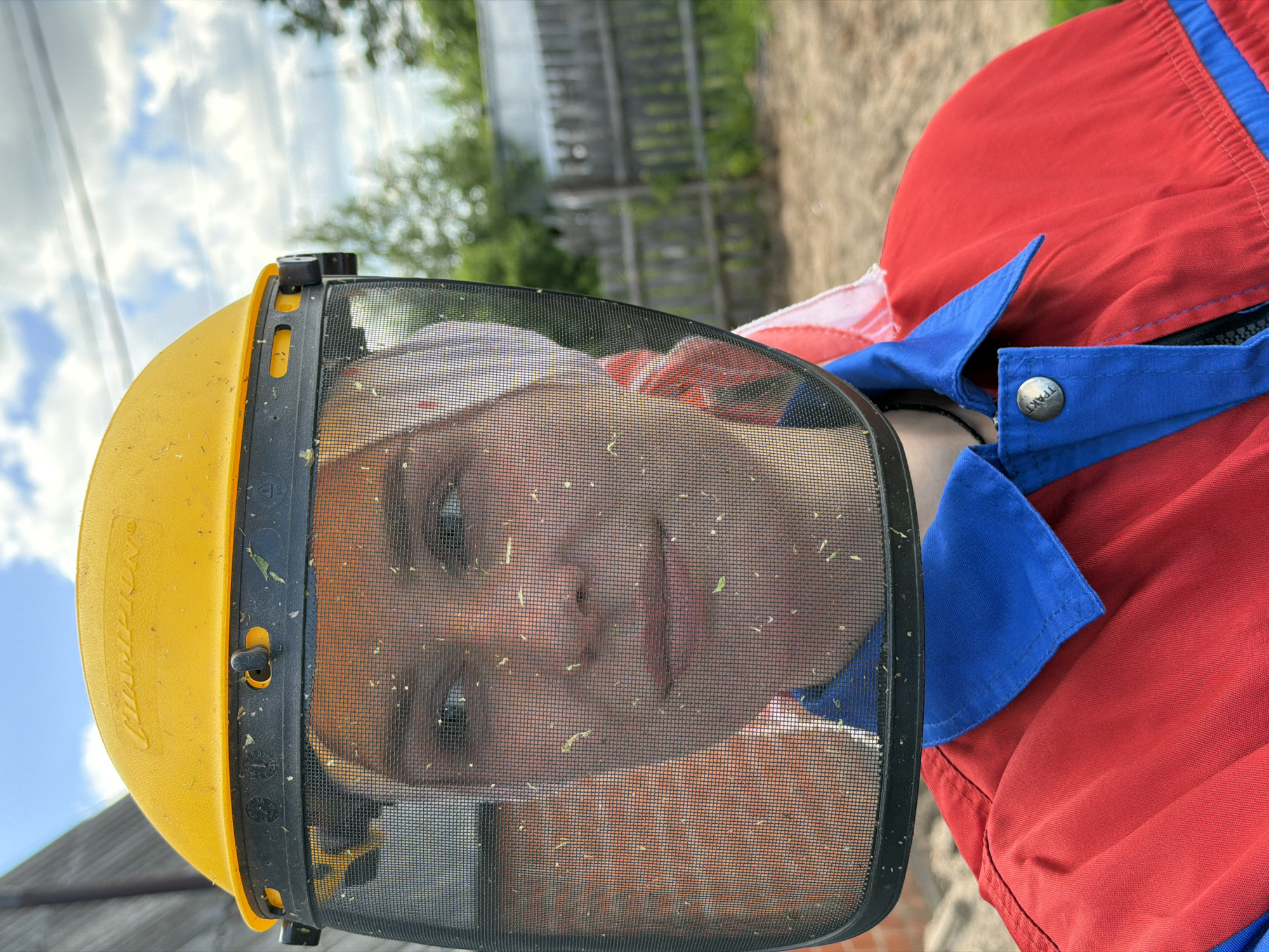 This is a close-up, vertical, outdoor shot of a person wearing a yellow hard hat and a mesh face shield. The face shield is dusty with small green flecks, likely grass or leaves, obscuring a portion of the person's face. The person's eyes are visible, looking forward, and their lips are slightly parted. They are wearing a red jacket with blue trim. A metal snap with the word "ТРАКТ" is visible on the blue trim of the jacket. The background shows a blurred wooden fence and green foliage, suggesting an outdoor, possibly rural or garden setting. The sky is visible at the top, with scattered clouds. The lighting suggests it is daytime. The scene is likely taking place in Chaikovsky, Russia.