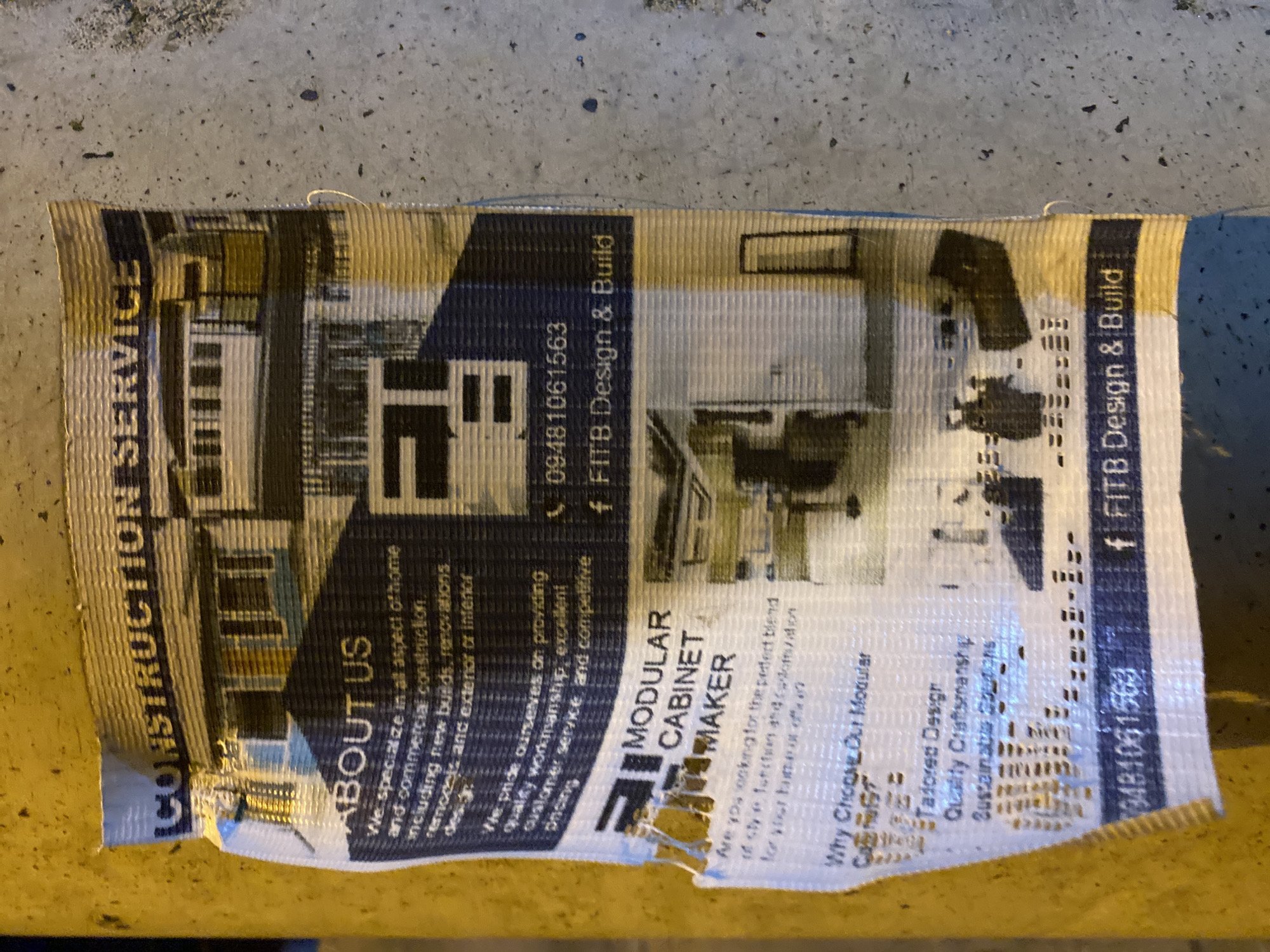 An outdoor advertisement flyer, significantly deteriorated, is affixed to a cylindrical yellow pole in Davao City, Philippines. The flyer, exhibiting multiple tears, creases, and extensive surface wear, displays information for a construction and design service.
The top portion of the flyer, horizontally aligned, reads "CONSTRUCTION SERVICE" above an illustration of a residential building. Below this, a section titled "ABOUT US" details the company's services, which include "home and commercial construction," "new builds, renovations, remodels," and "exterior or interior design." The text further states a commitment to "quality workmanship, excellent customer service and competitive pricing." A distinctive, dark-colored, angular logo, possibly representing "FITB," is positioned to the right of the "ABOUT US" text.
Beneath the "ABOUT US" section, a prominent heading reads "MODULAR CABINET MAKER." Accompanying text, partially obscured by tears and creasing on the left edge, asks, "Are you looking for the perfect blend of style, function and customization for your home or office?" This is followed by a section titled "Why Choose Our Modular Cabinets?", listing features such as "Tailored Design," "Quality Craftsmanship," and "Sustainable Solutions." Additional bullet points below are illegible due to severe damage. To the right of the cabinet maker details, images depict various modern kitchen or office cabinet installations.
Contact information is visible in two locations: midway down the flyer next to the logo, showing a phone icon followed by "09481061563" and a Facebook icon followed by "FITB Design & Build." The phone number and Facebook details are repeated at the bottom of the flyer, indicating "09481061563" and "f FITB Design & Build."
The flyer material appears to be a woven fabric or reinforced paper, displaying a subtle horizontal striped texture across its surface. The yellow pole to which it is attached suggests an urban or public infrastructure setting. The background consists of a rough, grey surface, consistent with concrete or asphalt, scattered with dark specks and minor debris.