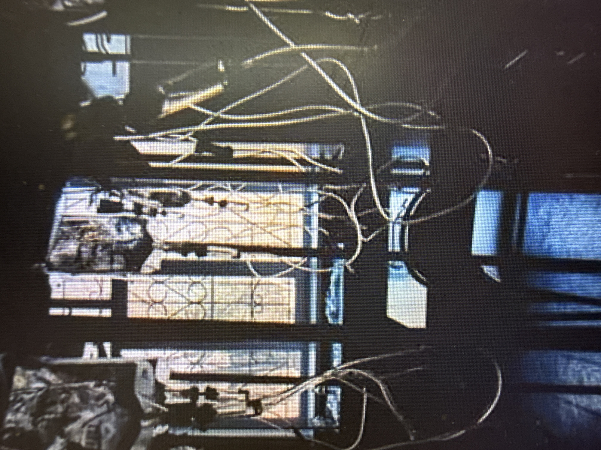 In an indoor setting, likely a room with a window featuring decorative metalwork, a series of clear plastic tubes is prominently displayed. These tubes are connected to what appear to be intravenous (IV) fluid bags suspended from above. The tubes snake across the scene, some coiled and others extending outwards, suggesting a medical or experimental context. The lighting is dim, with light filtering through the window, creating a somber atmosphere. The window itself is divided into panes, with an ornate pattern visible in some sections. The overall impression is one of a staged or controlled environment, possibly related to medical treatment, research, or a prop setup. The specific location within "Unknown, Unknown" is not discernible from the visual information.