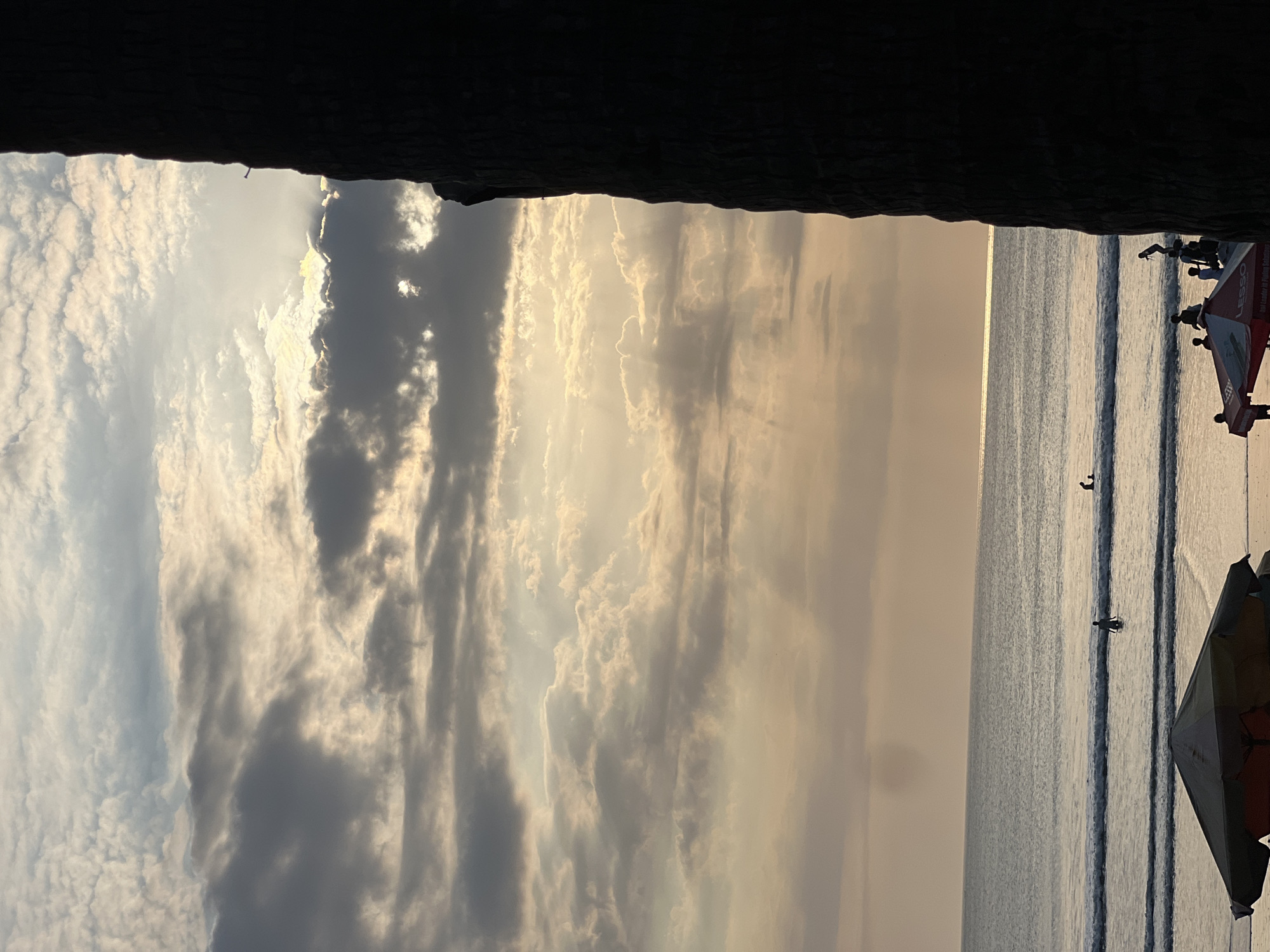 The image shows a beach scene at sunset in Kuta, Indonesia. The sky is filled with a dramatic display of clouds, with rays of sunlight breaking through, illuminating the scene with a warm, golden light. In the foreground, a dark, silhouetted tree trunk occupies the right side of the frame, adding a sense of depth and framing the view of the ocean and sky.
The ocean is calm, with gentle waves rolling towards the shore. A few people are visible in the water, some standing and others appearing to be swimming or playing. In the middle ground, two individuals are wading in the ocean. Closer to the shore, several people are gathered on the beach, some standing near umbrellas.
In the immediate foreground, a large, dark umbrella is positioned on the sand, partially obscuring the view of the beach. The overall atmosphere is serene and peaceful, capturing a typical evening on a tropical beach.