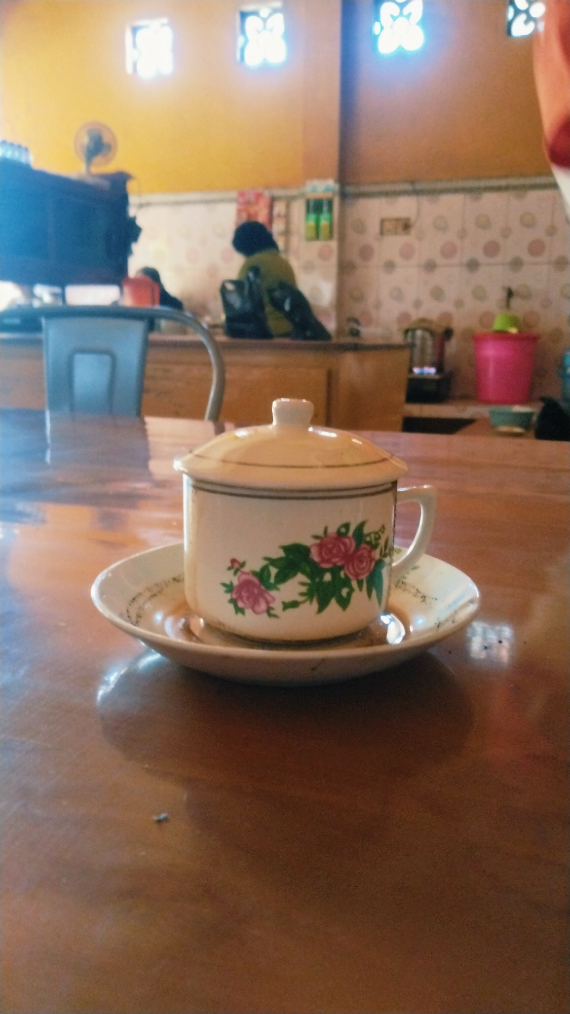 A coffee cup with a lid and floral decoration sits on a saucer on a wooden table. In the background, two people are visible, one sitting and facing away from the camera, and another partially obscured behind a dark cabinet. The room appears to be a cafe or eatery, with tiled walls and windows visible. The setting is consistent with the atmosphere of the city of Besuki, Indonesia, known for its local eateries and relaxed ambiance.