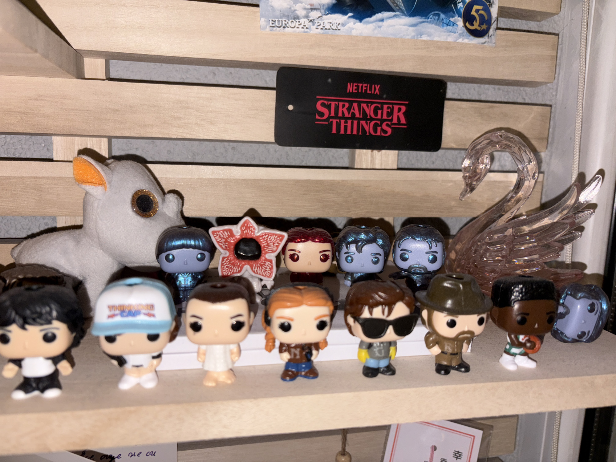 The image shows a collection of "Stranger Things" Funko Pop figures displayed on a wooden shelf. A gray stuffed animal is positioned behind the figures on the left. A crystal swan is on the right. A black "Netflix Stranger Things" sign is on the shelf behind the toys, as well as a "Europa Park" souvenir tag on the top shelf. The scene appears to be indoors, likely in a collector's home. The location in Panagyurishte, Bulgaria is unknown.