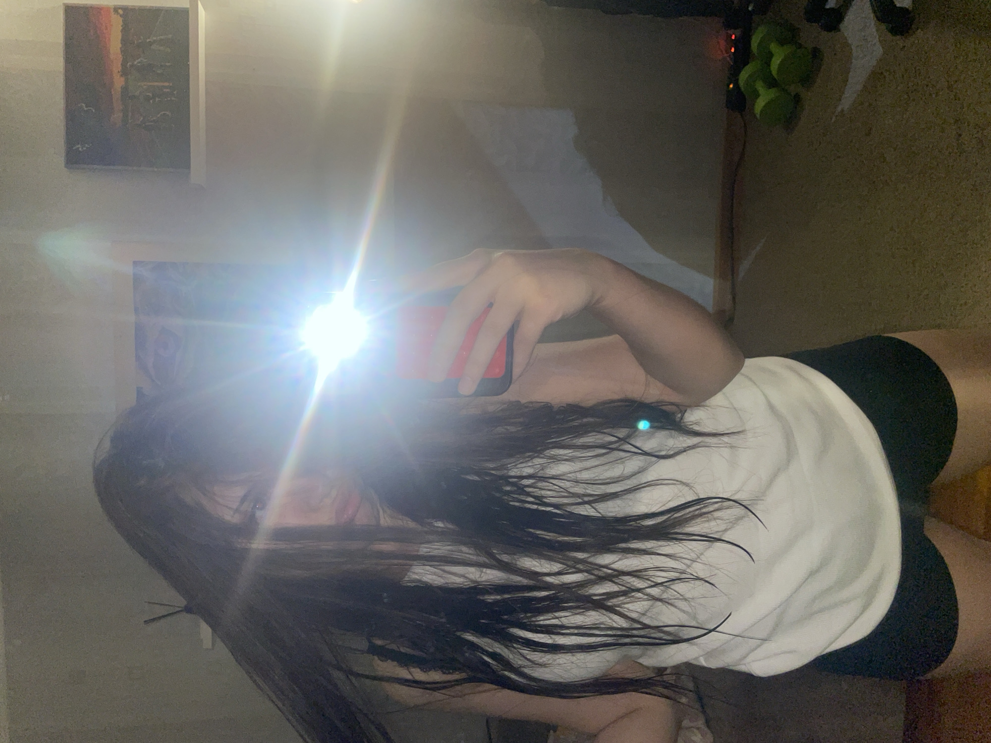 The image displays an indoor mirror selfie, rotated 90 degrees clockwise. A person, likely female, with long dark hair, holds a smartphone with a red case, using a bright flash that creates a significant glare, obscuring most of her face and upper body. She is wearing a white sleeveless top and dark bottoms, possibly shorts. Her hand and arm are visible, holding the phone.
In the background, on the left, a rectangular painting hangs on a light-colored wall, depicting a landscape with hues of orange and red, possibly a sunset or sunrise, and faint outlines of birds. Below it, another dark, less distinct picture or object is visible. To the right, some green, rounded objects, potentially small weights or decorative items, are arranged on a surface. The floor appears to be a dark, textured carpet or rug. The setting is an indoor room in Saint Petersburg, Russia, captured with a low-light flash, making specific time of day indistinguishable. No visible text is discernible in the image.
