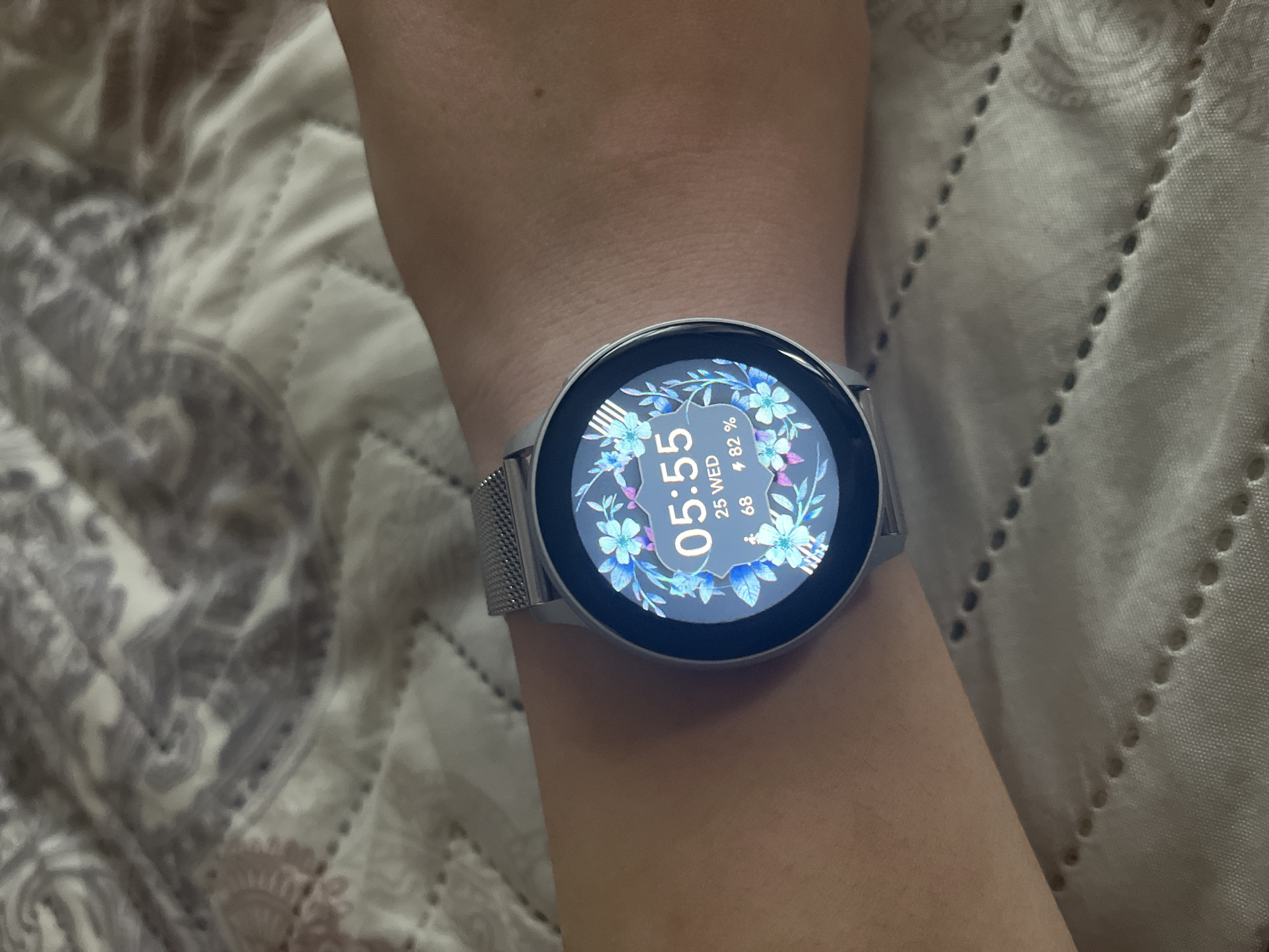A close-up photograph captures a human forearm and wrist, positioned against a light-colored, quilted fabric background. A silver-toned smartwatch, equipped with a silver mesh band, is worn on the wrist. The circular watch display is active, showing the digital time as 05:55, the date as 25 WED, a heart rate of 68 beats per minute, and a battery charge of 82%. The watch face features a decorative border composed of blue and purple floral elements. The background fabric exhibits a faint, repeating pattern in muted grey and off-white tones, with discernible rows of stitching forming raised dots. The scene is static and takes place indoors, potentially within the city of Varna, Bulgaria.
