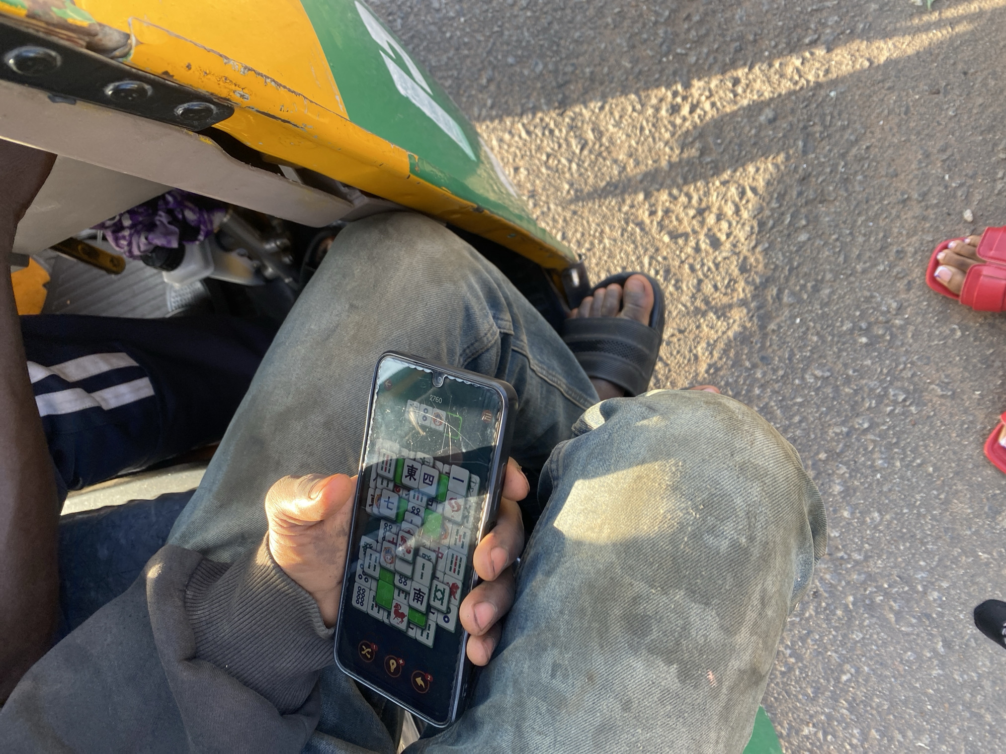 In Jos, Nigeria, a person is seated in what appears to be a motorized tricycle, commonly known as a "Keke" or "Marwa." The individual is wearing blue jeans and a grey sweatshirt, with their left hand holding a smartphone. The phone displays a game that resembles Mahjong, with various tiles visible. The person is also wearing black sandals. In the background, a portion of the yellow and green Keke is visible, along with what looks like a purple fabric object. The ground is paved with asphalt, illuminated by sunlight casting shadows. Another person's feet, wearing red sandals, are visible in the lower right corner of the image. The scene captures a moment of casual engagement with a mobile game while possibly traveling in a Keke in Jos.