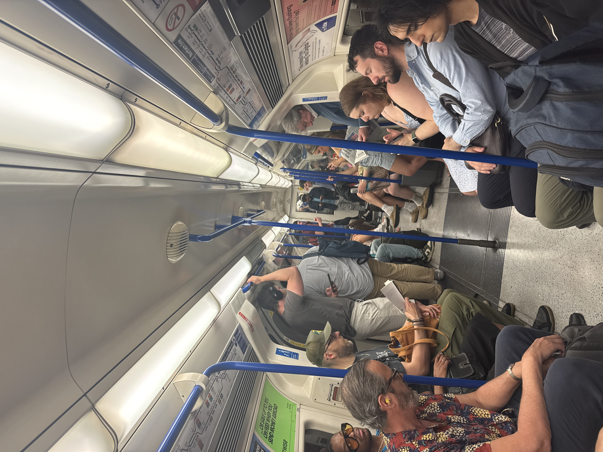 The image captures the interior of a crowded subway train, likely in London, based on the visible "Northern Line" signage and the "Tide" advertising. Passengers are seated and standing close together, many focused on their phones, books, or personal space. The interior of the train features white panels, blue handrails, and advertising posters. The lighting is bright, creating a somewhat clinical atmosphere. People are diverse in age, dress, and ethnicity.