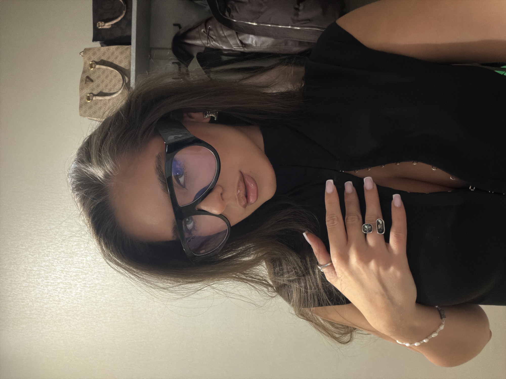 A woman with long brown hair, wearing black-framed glasses and a black low-cut top, is seen in a close-up, slightly angled shot. She is wearing two rings on her right hand and a bracelet. Behind her, partially visible are handbags placed on shelves, suggesting an indoor environment, potentially a closet or a store. The location in Krasnoyarsk, Russia is not discernible from the image.