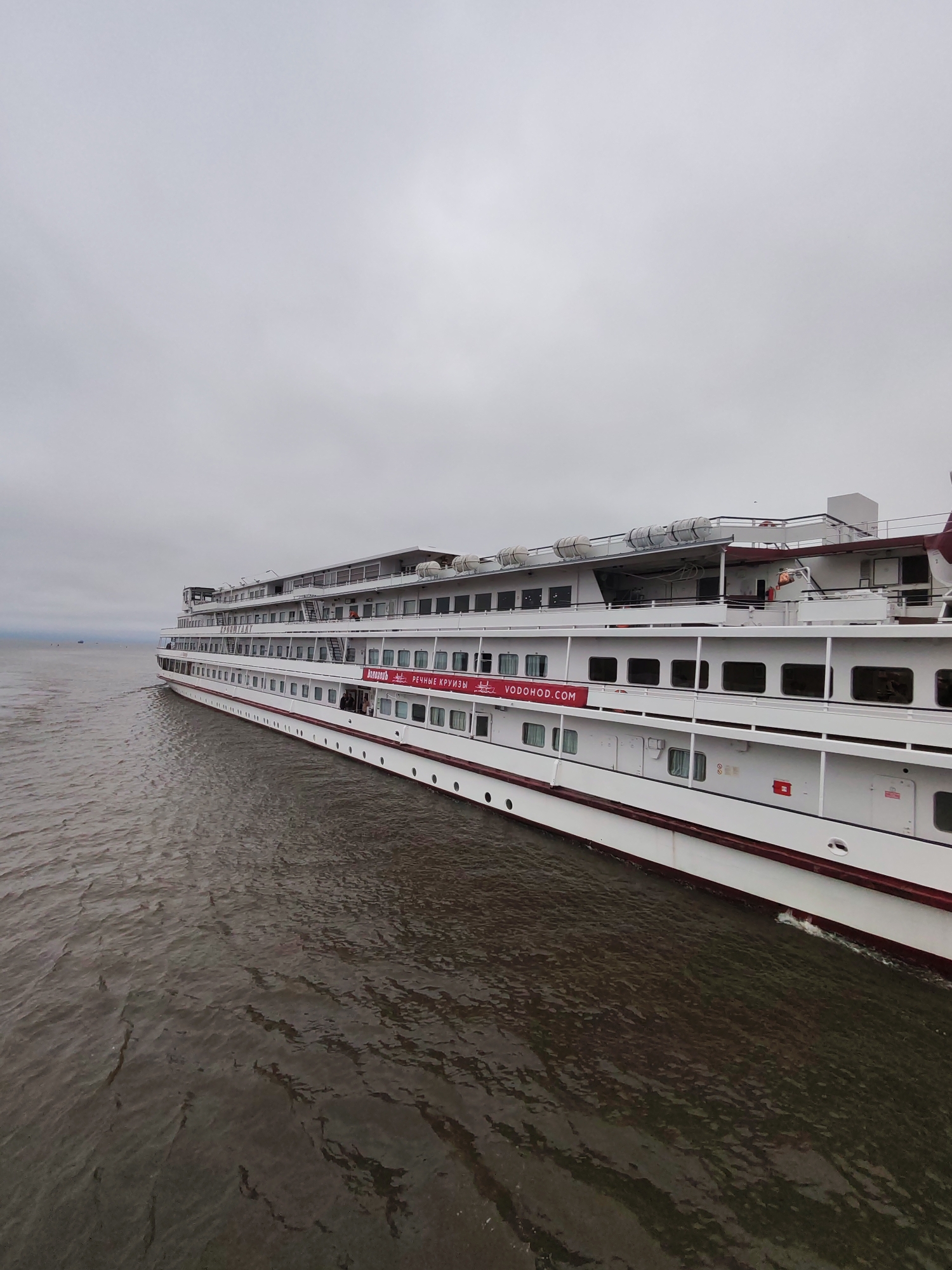 A white cruise ship with red trim is sailing on dark, choppy water under a cloudy sky. The ship has multiple decks with many windows. A red banner with white text that reads "РЕЧНЫЕ КРУИЗЫ VODOHOD.COM" is visible on the side of the ship. There are several life rafts visible on the top deck. The ship appears to be in motion, with a slight wake visible in the water. The location is the city of Krokhino, Russia.