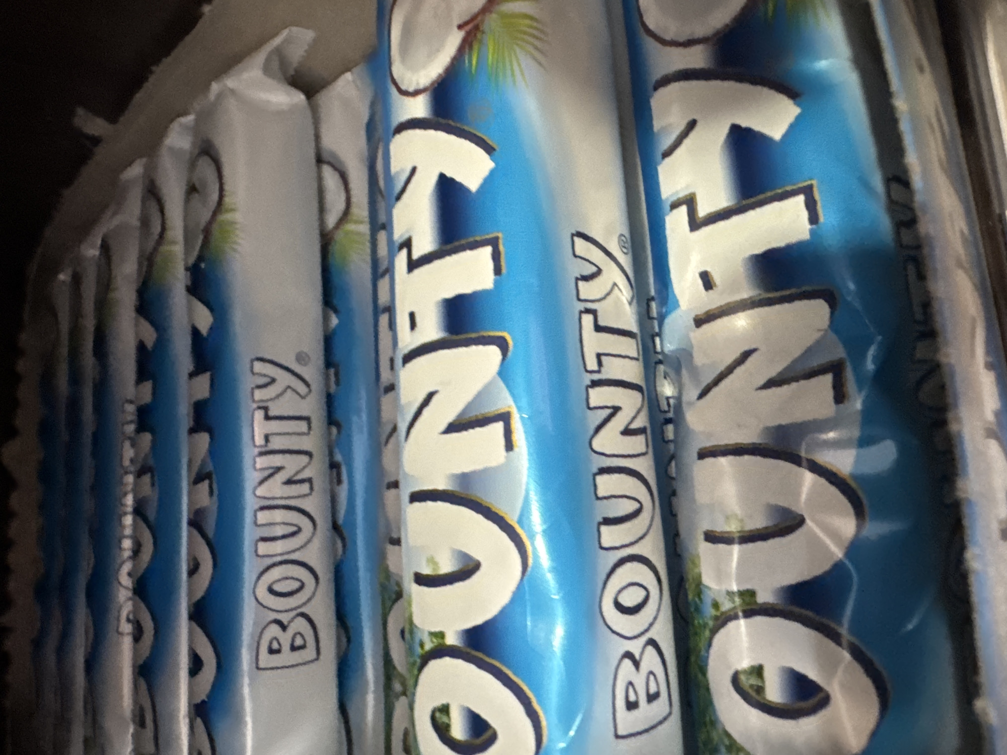 The image presents a close-up view of multiple Bounty chocolate bars, arranged vertically in what appears to be a display or storage setting. The bars are individually wrapped in distinctive blue and white plastic packaging.
Each wrapper prominently features the brand name "BOUNTY" in large, white, sans-serif lettering with a dark blue outline, set against a bright blue background. Many of the wrappers also display an illustration of a cracked coconut with green fronds, indicating the coconut flavor of the product. The overall design of the packaging is consistent across all visible bars.
The bars are stacked or placed in rows, with their narrow sides facing the camera. The background behind the products is dark and out of focus, suggesting an indoor environment such as a shop shelf or a storage area. No people are visible, and there is no specific activity occurring; the image is a static shot focused entirely on the packaged food items. The lighting is uniform, highlighting the product details.
