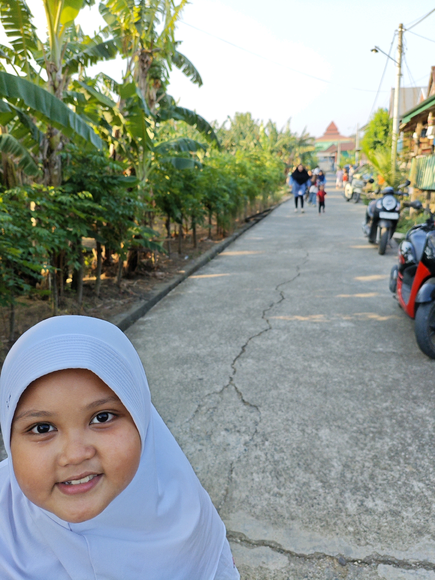 In Padduppa, Indonesia, a young girl wearing a white hijab is the foreground subject of this image. She is looking up and smiling at the camera. Behind her, a paved road extends into the distance, lined with banana trees and other lush vegetation on the left. On the right side of the road, several motorcycles are parked. Further down the road, a group of people, including adults and children, are walking away from the camera. In the background, buildings and a structure with a red roof are visible under a bright, clear sky. The scene appears to be a village or rural setting during the day.