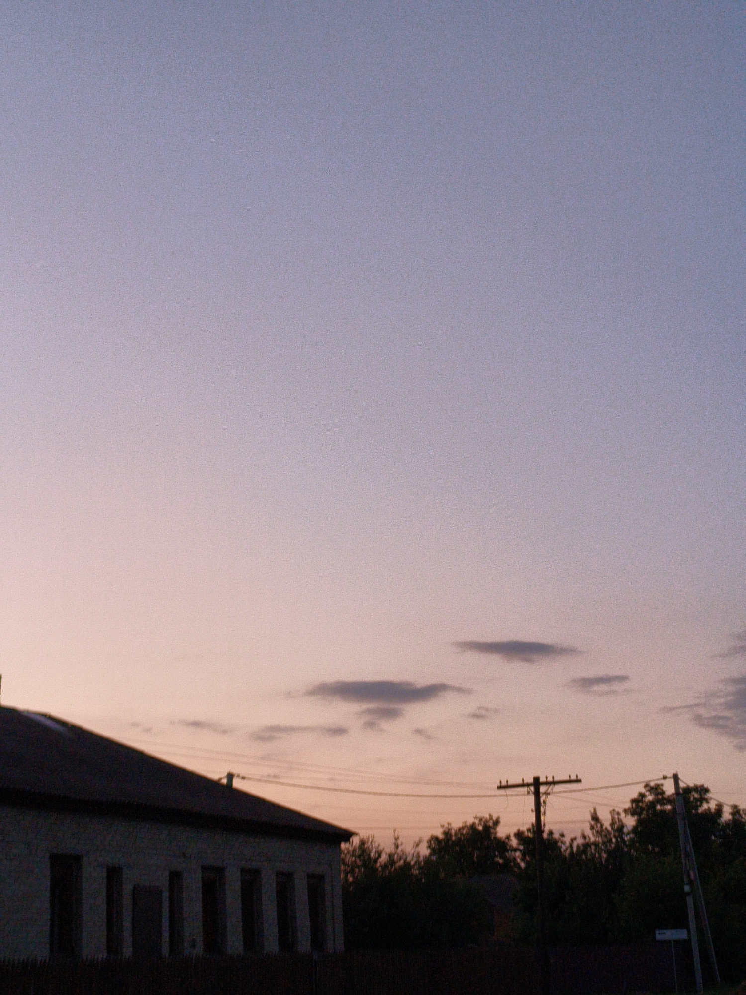 The image depicts a twilight scene in Ustyvytsia, Ukraine. In the foreground, a long, single-story building with a dark, sloped roof is visible on the left. The building's exterior appears to be made of stone or concrete, and it has several dark, recessed doorways or windows. A faint outline of trees can be seen along the right edge of the building.
The middle ground of the image features a rural landscape. A wooden utility pole with crossbars and insulators stands prominently, supporting power lines that stretch across the frame. Another utility pole, appearing to be made of concrete or metal, is partially visible on the far right, also with associated wires. Behind these poles, a dense line of dark green trees forms a natural barrier. The silhouette of another, smaller building is subtly visible amongst the trees.
The background is dominated by a serene sky, transitioning from a soft lavender at the top to a pale pink near the horizon. A few wispy, dark clouds are scattered across the sky, adding depth to the scene. The overall atmosphere is calm and quiet, typical of a rural evening in Ustyvytsia. There are no people or animals visible in the image.