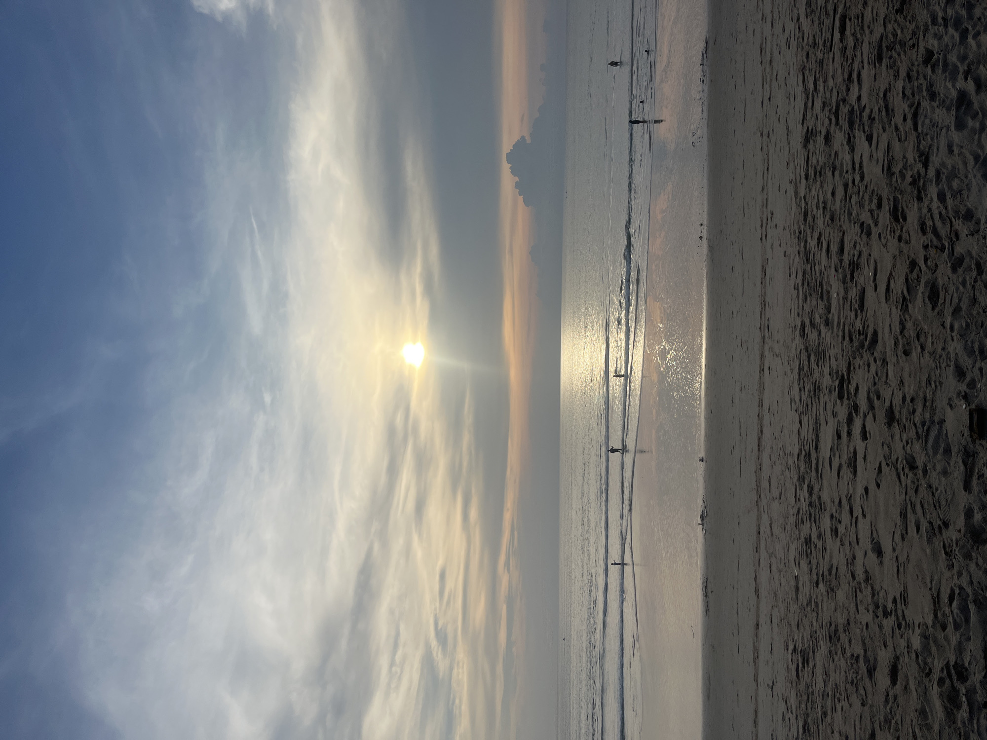 This is a vertical, eye-level, medium shot of a tranquil beach scene at sunset in Choeng Thale, Thailand. The sun, a bright, slightly hazy orb, is low in the sky, casting a golden glow across the water and the sand. Wispy clouds streak across a blue and pale orange sky. The ocean stretches out to the horizon, with gentle waves lapping the shore. Several small figures, appearing as silhouettes, are scattered along the water's edge, some wading, others standing. The wet sand reflects the sky and the sunlight, creating a shimmering surface. The foreground is dominated by dry, dark sand, textured with footprints. The overall atmosphere is peaceful and serene, capturing the end of a day at the beach.