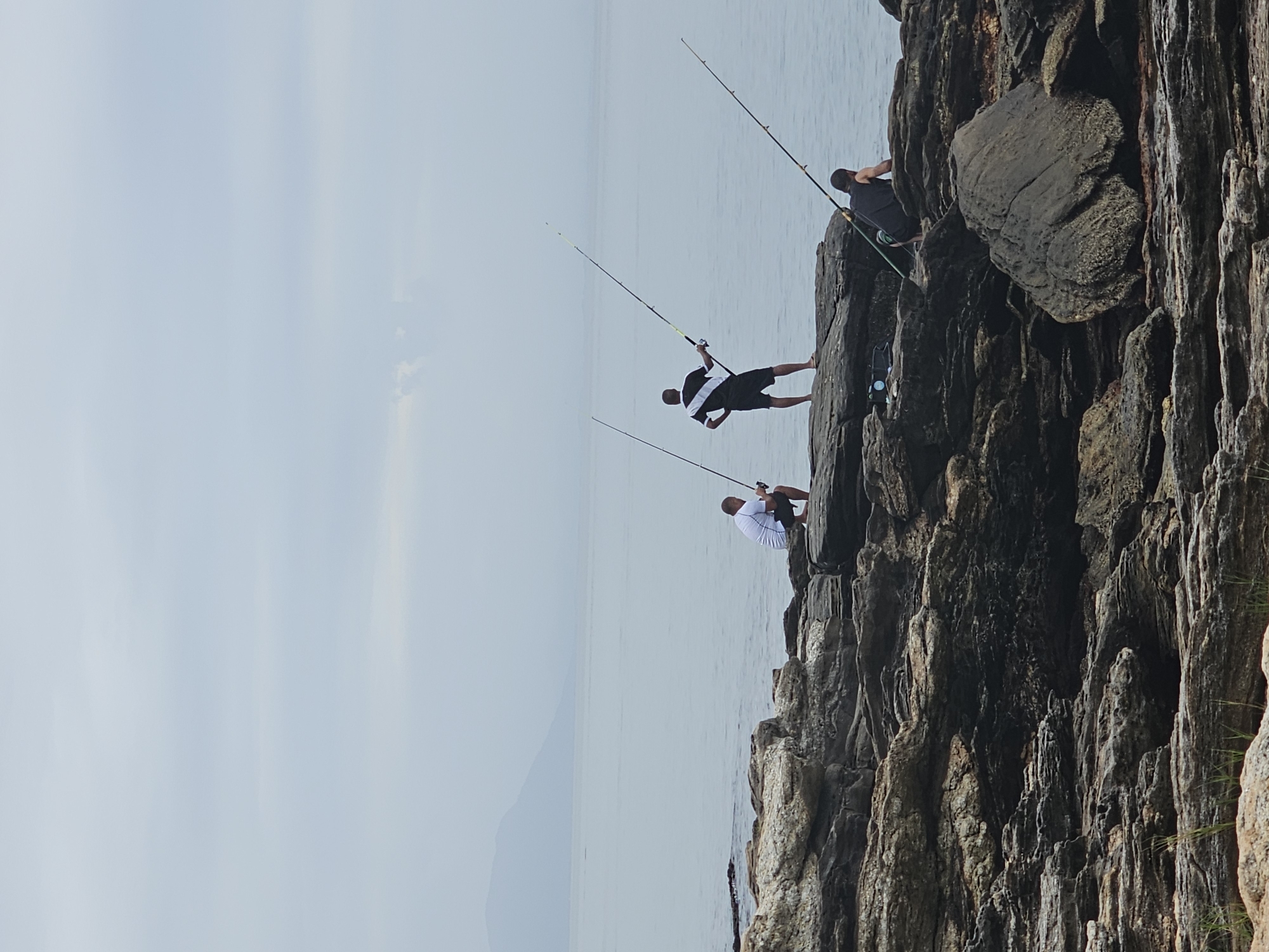 Three men are fishing from rocky cliffs in Armação dos Búzios, Brazil. The men are positioned along the edge of the rocks, each holding a fishing rod. The rods are arched, indicating they are casting or waiting for a bite. The sea is visible in the background, along with a faint outline of a mountain on the horizon. The sky is overcast. The rocks are dark and textured.