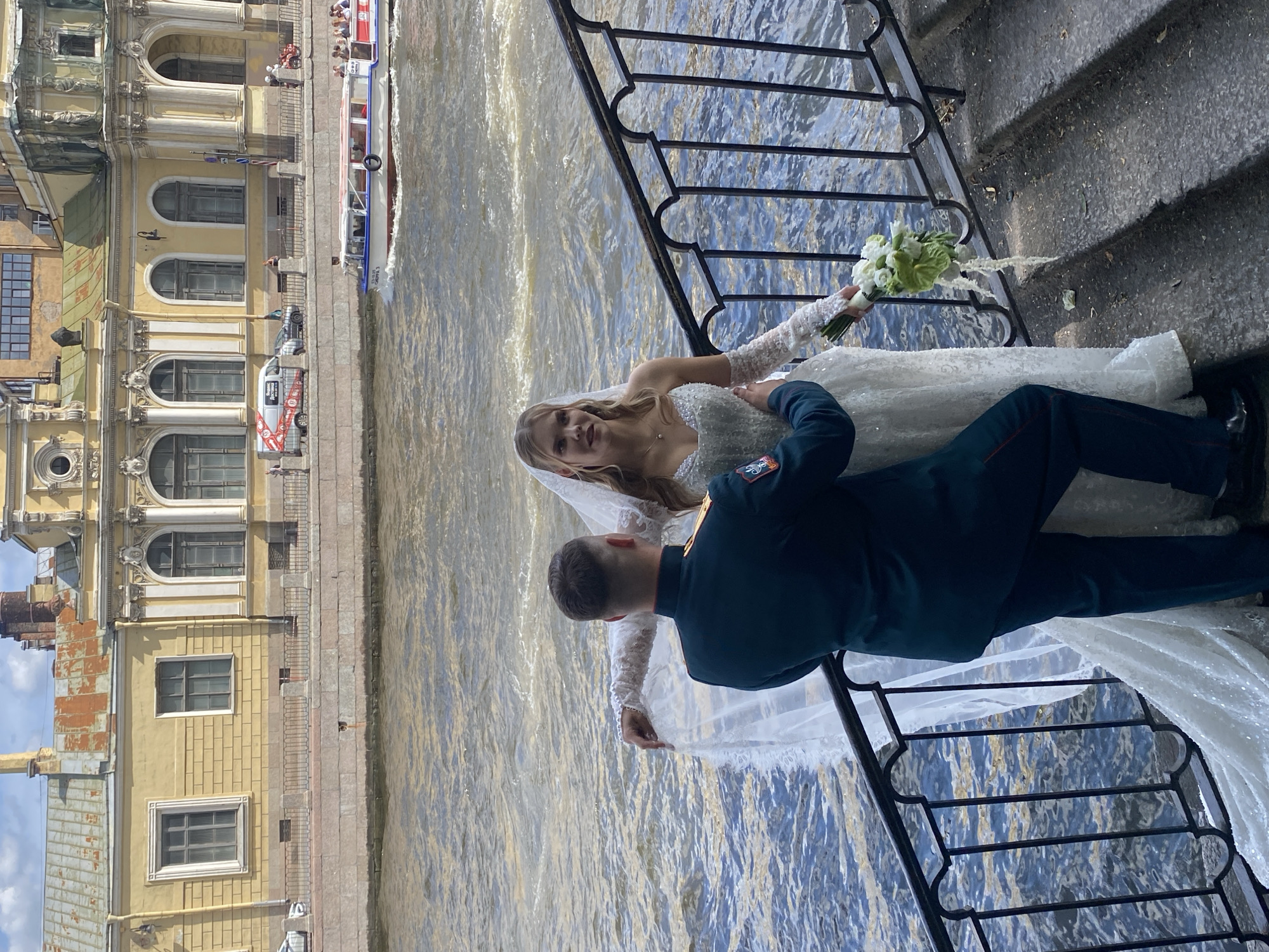 A newly married couple is posed on a stone staircase next to a railing, with a river and a yellow building in the background. The groom, dressed in a dark green military uniform with red stripes on his trousers, is seen from behind, his left arm around the bride. The bride, wearing a white, sparkling wedding dress with long lace sleeves and a veil, faces the camera. She holds a bouquet of white and green flowers. The river behind them is choppy, and a white tour boat with a red roof is visible in the distance. The building in the background is elaborate with classical architectural details and multiple windows. The overall scene suggests a wedding photoshoot in Saint Petersburg, Russia, likely along the Neva River or one of its canals.