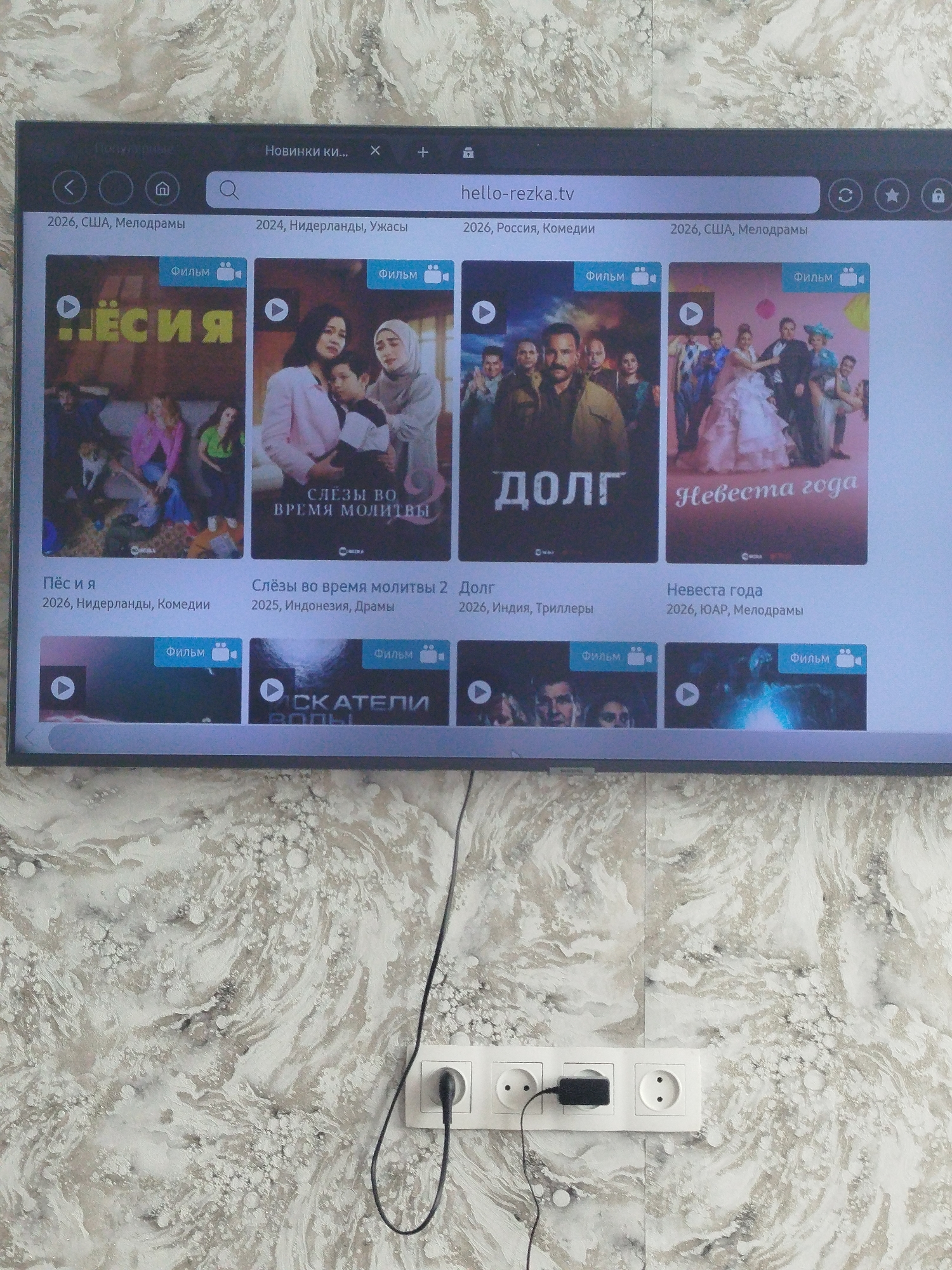 The image shows a television screen displaying a website with movie titles and posters. The website is likely a streaming service, as indicated by the presence of movie thumbnails, titles, years, genres, and play buttons. The screen displays a browser interface with a search bar that contains the text "hello-rezka.tv". Below the browser interface, there are several movie posters with accompanying text. Some of the visible movie titles include "Лёсия", "Слезы во время молитвы", "Долг", and "Невеста года". The genres listed include "Мелодрамы", "Комедии", "Ужасы", and "Триллеры". The screen is mounted on a wall with a distinctive patterned wallpaper. In the lower part of the image, a white power strip with three outlets is visible, with a black power cord plugged into one of them, and another black plug inserted into an outlet on the wall. This suggests a domestic setting. The location of Залескі сельскі Савет, Belarus, is not visually discernible from the provided image.