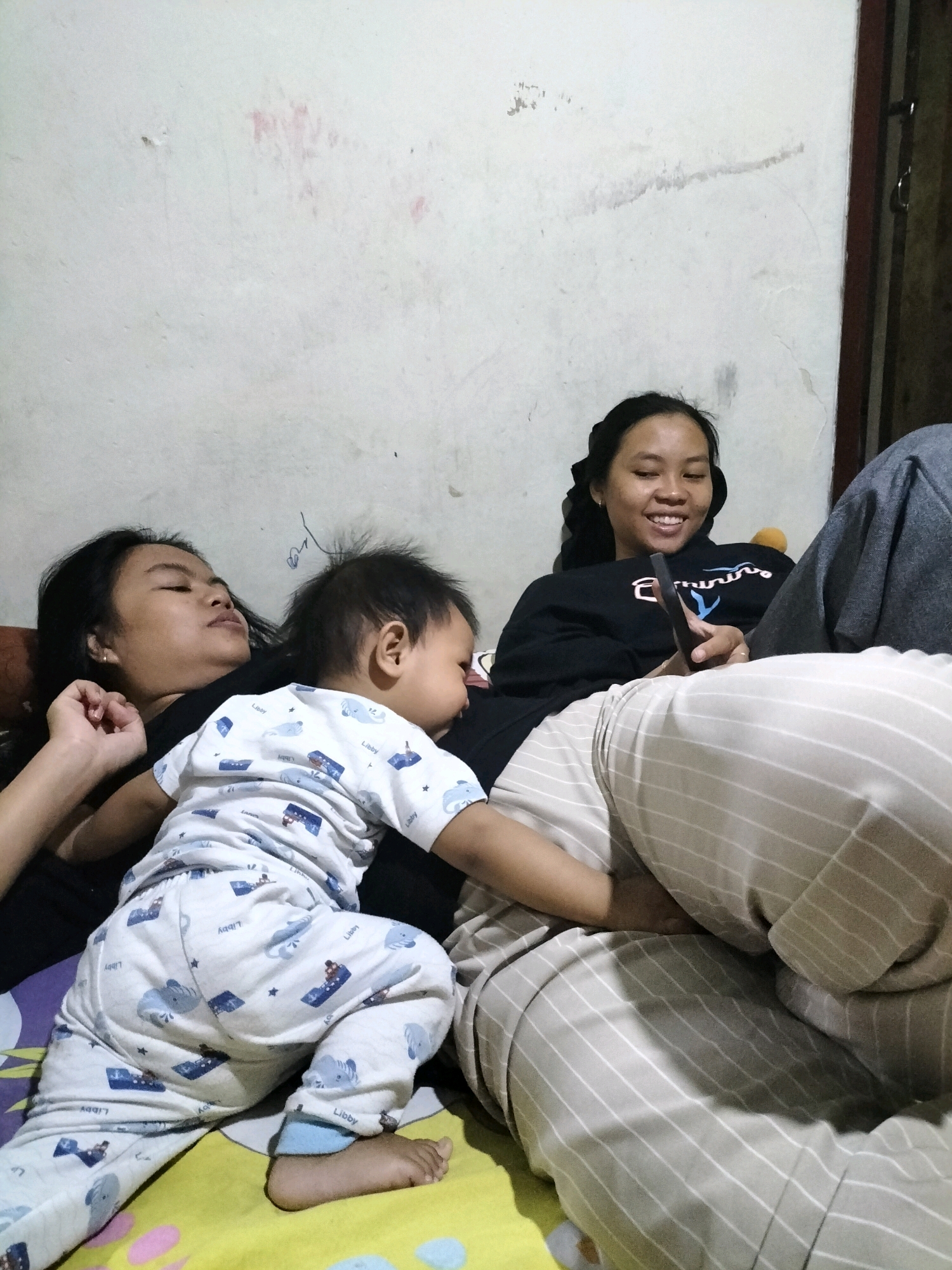 In a domestic interior setting, likely a bedroom in Tasikmadu, Indonesia, three individuals are depicted lying down. A young child, dressed in white pajamas with a pattern of blue elephants and the word "Libby," rests on the chest of an adult. The child's face is turned away, and its left arm is extended, resting on the adult's shoulder. To the right, another adult is reclined, holding a smartphone in their right hand and smiling while looking at it. This adult is wearing a black hoodie with white lettering and appears to be observing content on the device. The background consists of a textured, off-white wall with some faint stains and marks, and a dark wooden door frame is partially visible on the right. The individuals are situated on a surface covered with a yellow and green patterned sheet.