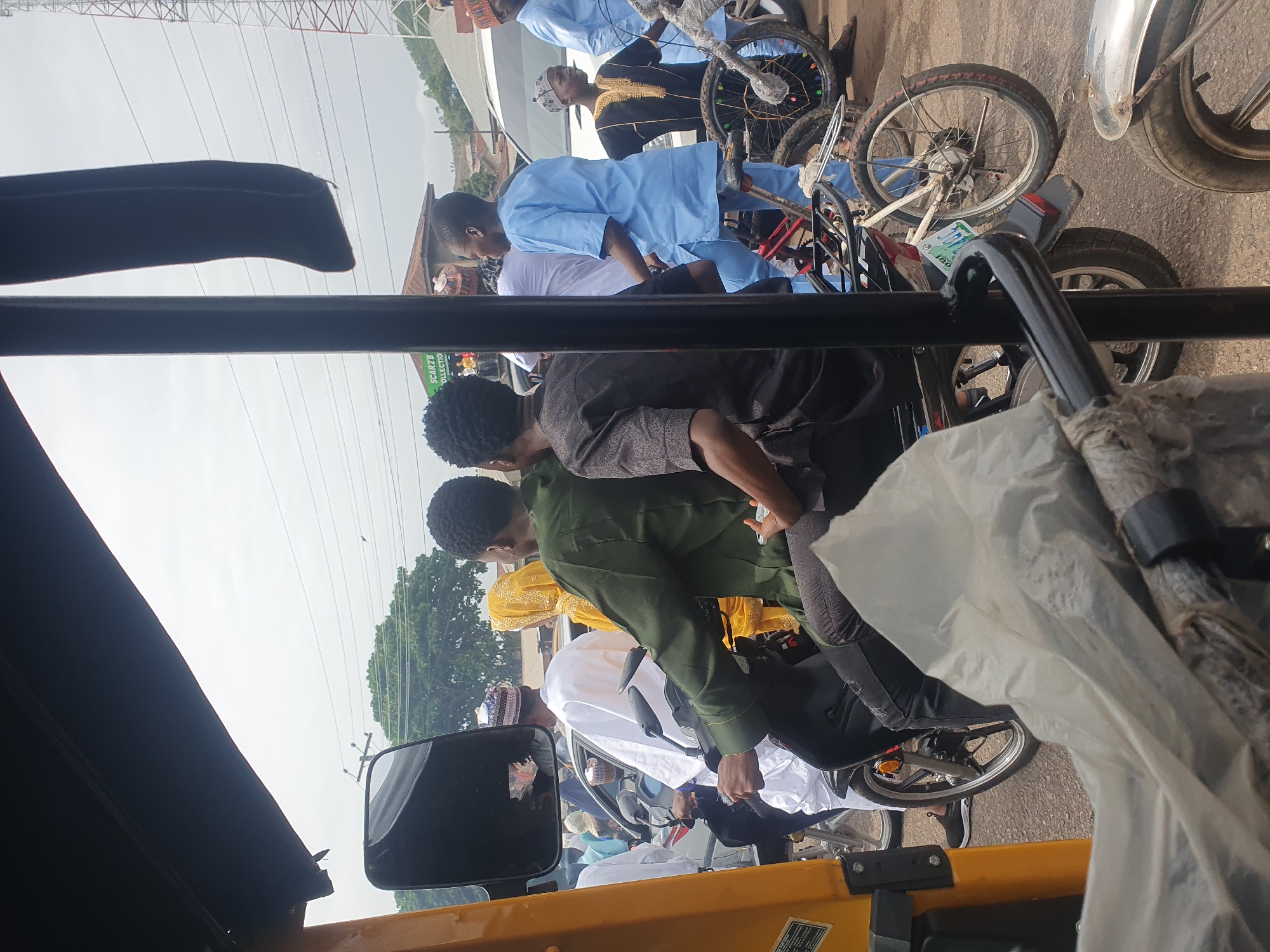 The image, taken from the interior of a yellow motorized tricycle, shows a street scene in Lokoja, Nigeria. A group of people, several in traditional clothing, are standing near or riding bicycles. Some individuals wear light blue robes while others wear more ornate attire. The view is partially obstructed by the tricycle's frame and side mirror. Overhead power lines are visible against a bright sky.