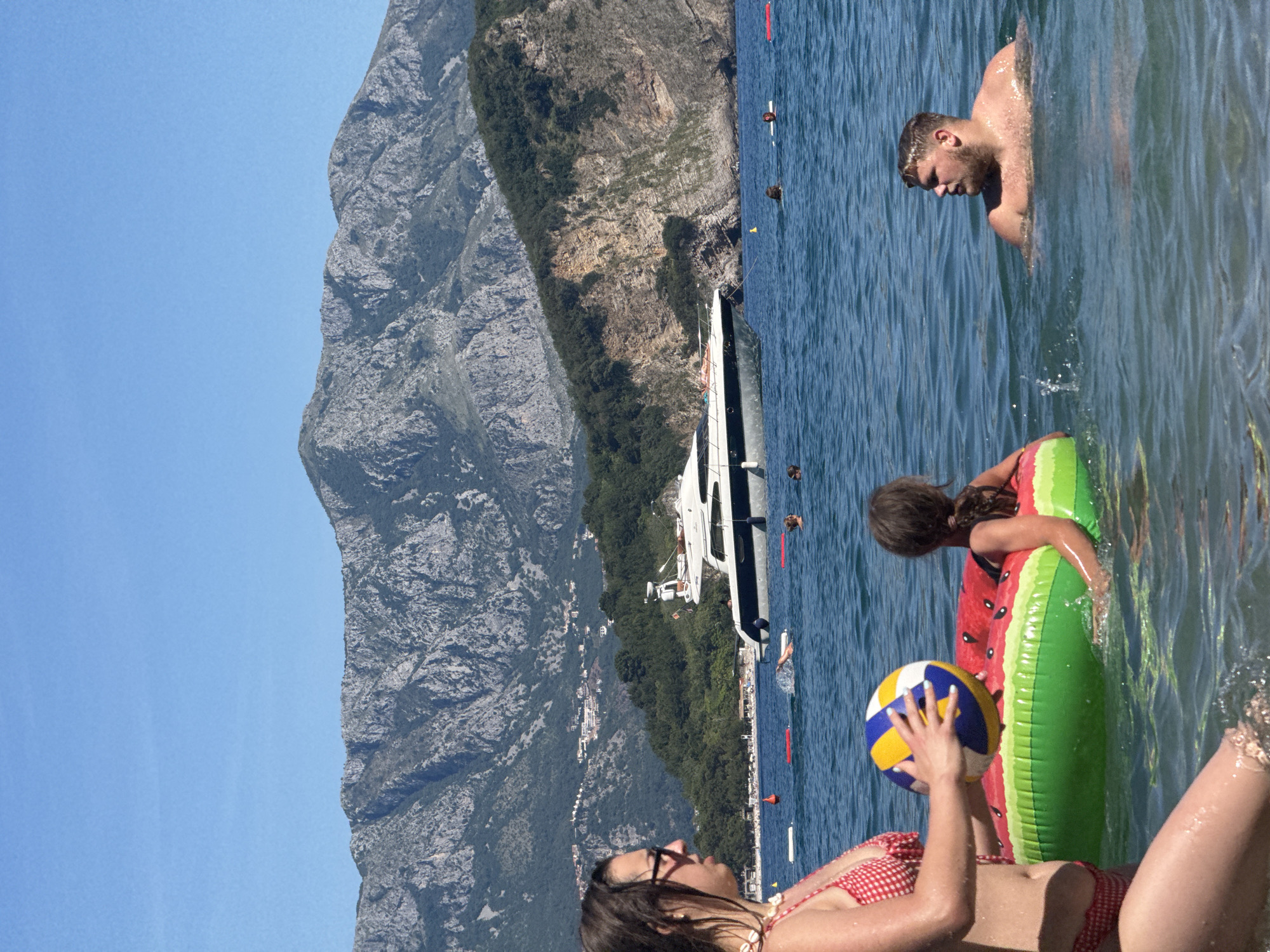 The image depicts a sunny day at a beach in Budva, Montenegro. In the foreground, a woman in a red and white checkered bikini holds a volleyball. Beside her, a young girl floats in a watermelon-shaped inflatable ring. To the right, a man with blond hair is partially submerged in the water, looking to his right. In the mid-ground, a white yacht with a dark stripe is anchored in the blue sea. Behind the yacht, a rocky mountainside covered with green vegetation rises, with buildings visible on its slopes. The sky is clear and blue. The scene suggests a leisurely day at the beach with recreational activities in the water.