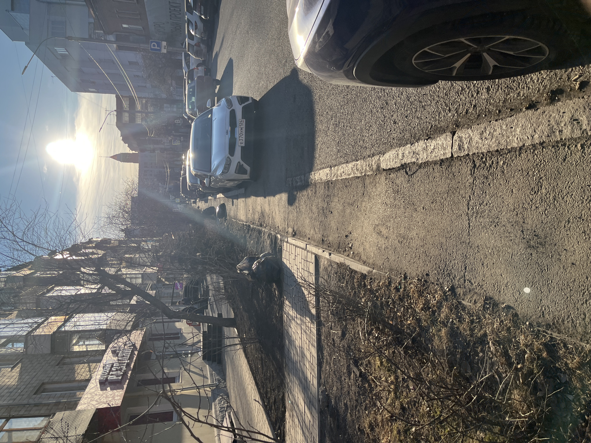 The image shows a street scene in an urban area. Cars are parked along the right side of the street, with a white Kia being prominently visible. The street is paved and has a white line marking the edge. A sidewalk runs alongside the street, bordered by a patch of dry vegetation. Buildings are visible on both sides of the street, with some featuring architectural details. The sun is shining brightly in the distance, casting long shadows. A tower-like structure can be seen in the background between the buildings. A sign with lettering in Russian is visible on one of the buildings to the left. The license plate of the white car reads E370YM124.