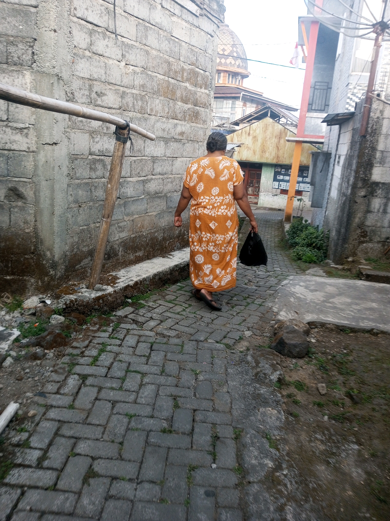 The image depicts a woman walking down a narrow, brick-paved alleyway in Andongsili, Indonesia. She is wearing a long, orange dress with a white floral pattern and carrying a black plastic bag in her right hand. The alley is flanked by rough, concrete block walls on both sides. To the left, a rustic bamboo pole is propped against the wall. In the background, several buildings are visible, including one with a domed roof that suggests a religious structure. The overall scene portrays a typical, modest urban environment.