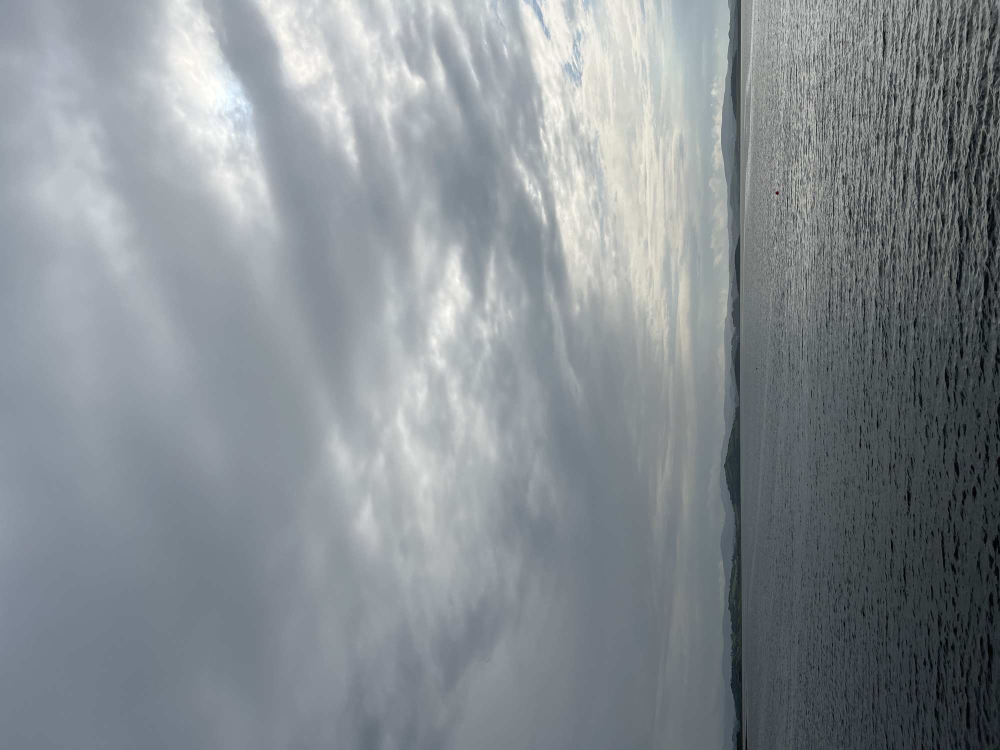 A wide shot captures a body of water against a backdrop of a cloudy sky and distant hills. The water, dark and textured with ripples, occupies the lower half of the frame. A small, red buoy is visible in the mid-right section of the water. The horizon line separates the water from the sky and land. The sky is filled with various shades of grey clouds, with some brighter patches suggesting sunlight breaking through in the upper right. The distant hills are rendered in muted, hazy tones, with a few faint structures possibly visible on their slopes. The scene appears to be taken from a vantage point on the shore or a vessel, overlooking a bay or inlet. The overall mood is somewhat overcast and serene. The image is likely taken in or around Mountcharles, Ireland, given the presence of hills and coastal waters characteristic of the area.
