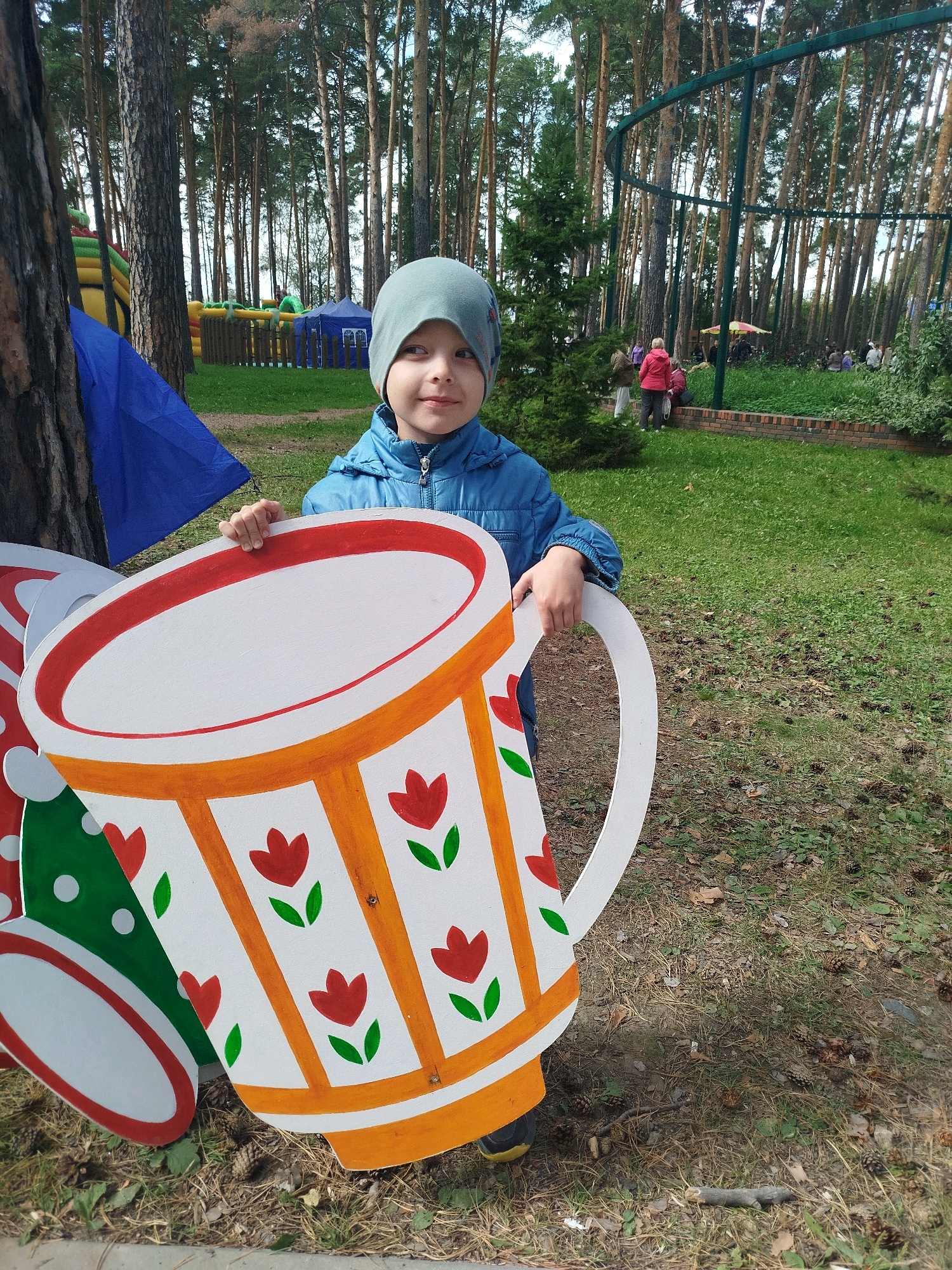 A young child, likely a boy, wearing a light blue jacket and a grey beanie, stands in a park, holding a large, colorful cutout of a teacup. The teacup cutout is white with orange stripes and red floral designs. In the background, several tall pine trees create a dense forest. Further in the distance, there are people gathered in what appears to be a recreational area with bouncy castles and tents, indicating it's likely a public park or an event space. The ground is covered with grass and fallen pine needles. The child is looking towards the camera with a slight smile. The scene suggests a daytime setting, possibly during a warm season. The location is specified as Zavodoukovsk, Russia.