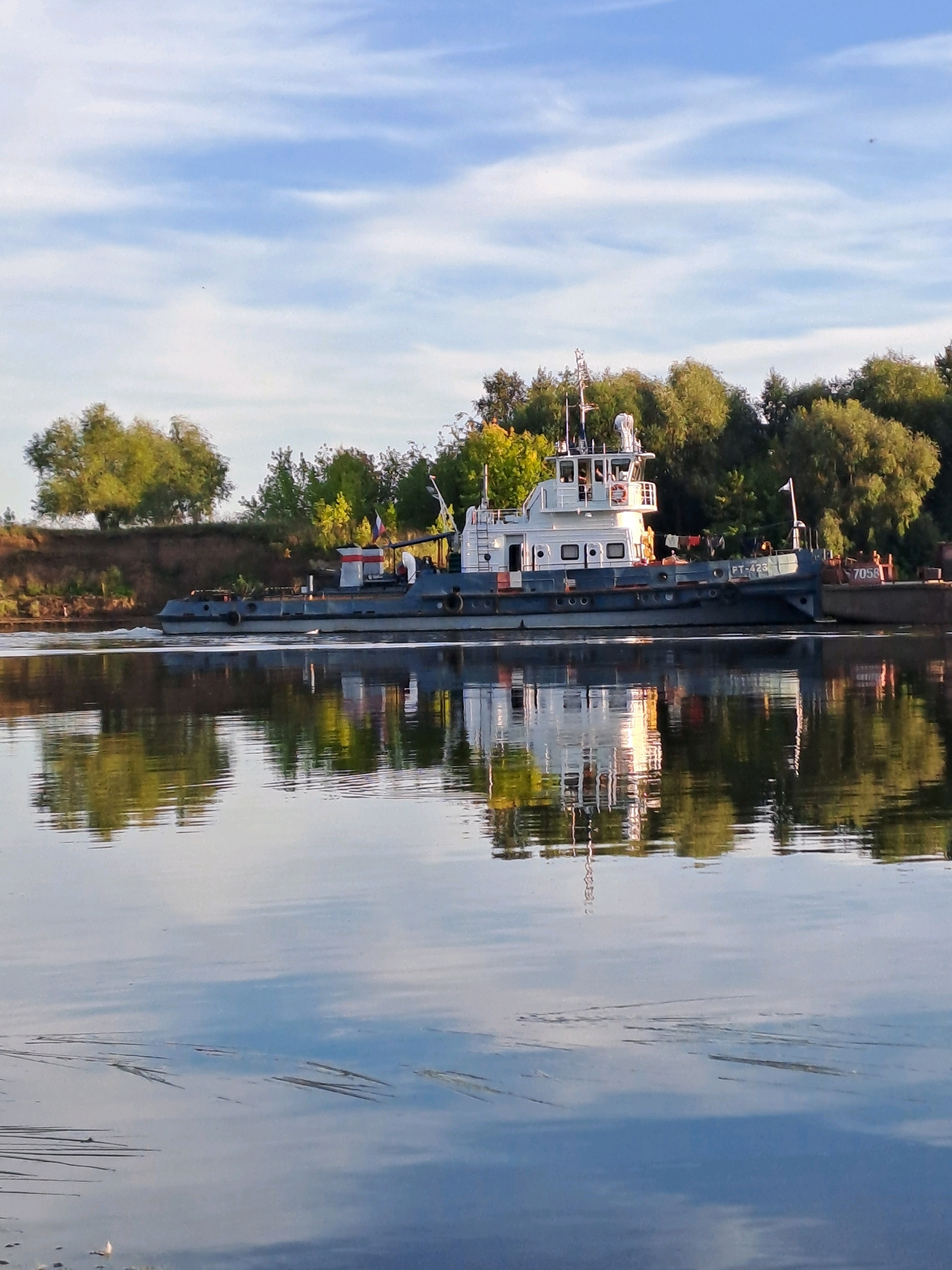 A tugboat, identified by the markings "PT-426" and "7058", is depicted in motion on a calm river under a blue sky with scattered clouds. The tugboat, predominantly white and dark blue, has a superstructure with multiple levels and windows, and a mast with equipment. Two flags, one red and white and another white with a red symbol, are visible. The tugboat appears to be pushing or towing a barge to its right. The river's surface reflects the tugboat, the barge, and the lush green trees lining the riverbank. The background consists of a sloped embankment covered in dense foliage. The scene appears to be a serene natural environment. The location is the city of Beloomut, Russia, suggesting a body of water such as the Oka River.