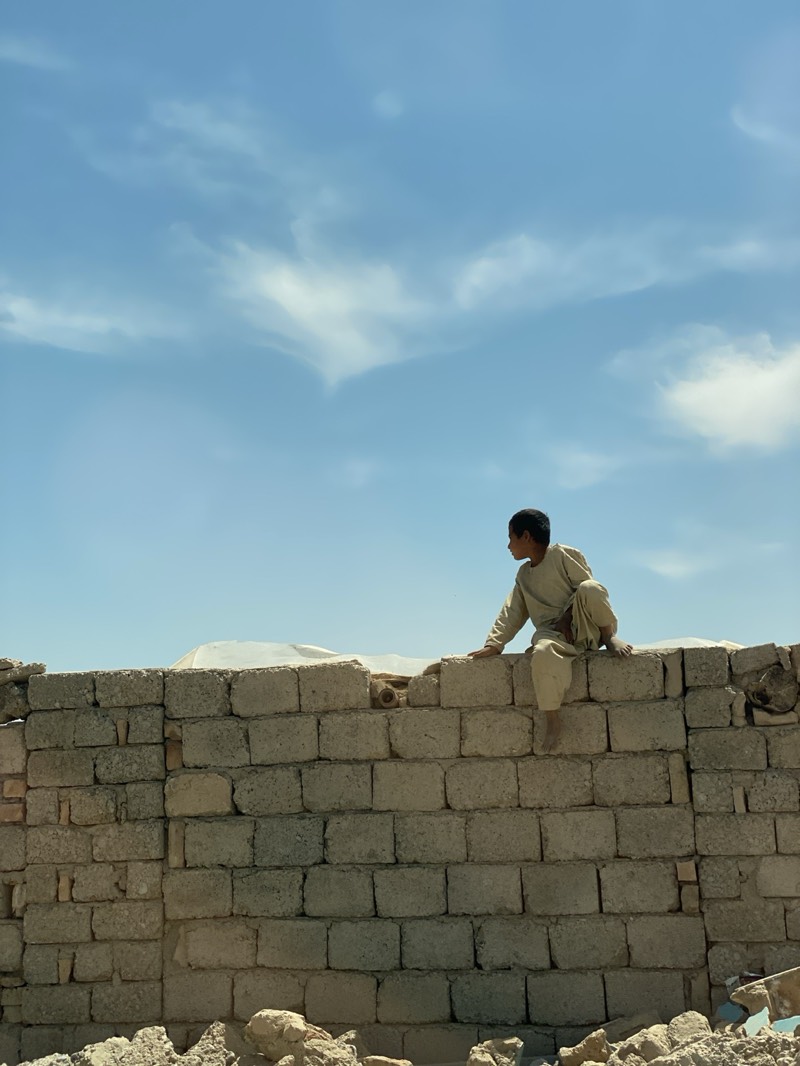 A young boy is sitting on top of a brick wall, with a cloudy sky in the background. The wall appears to be made of light-colored, rectangular bricks. The boy has dark hair and is wearing a light-colored outfit. In the background, there is a white tarp. The photo was taken in Deh-E Bagh, Afghanistan.