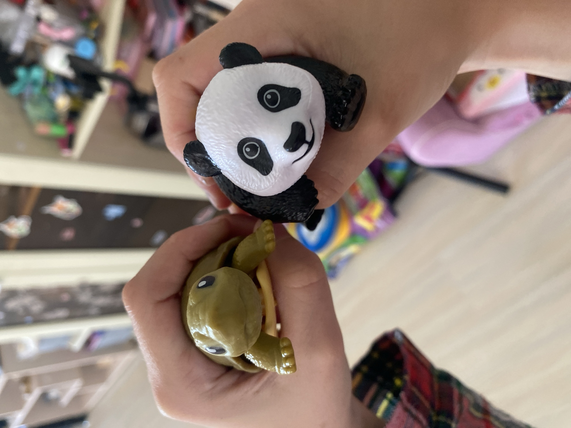 Two hands are holding two toy animals. The left hand is holding a green toy turtle. The right hand is holding a black and white toy panda. The background is blurred, but appears to be an indoor setting with shelves and toys. There is no indication of the location being the city of Volzhsk, Russia.
