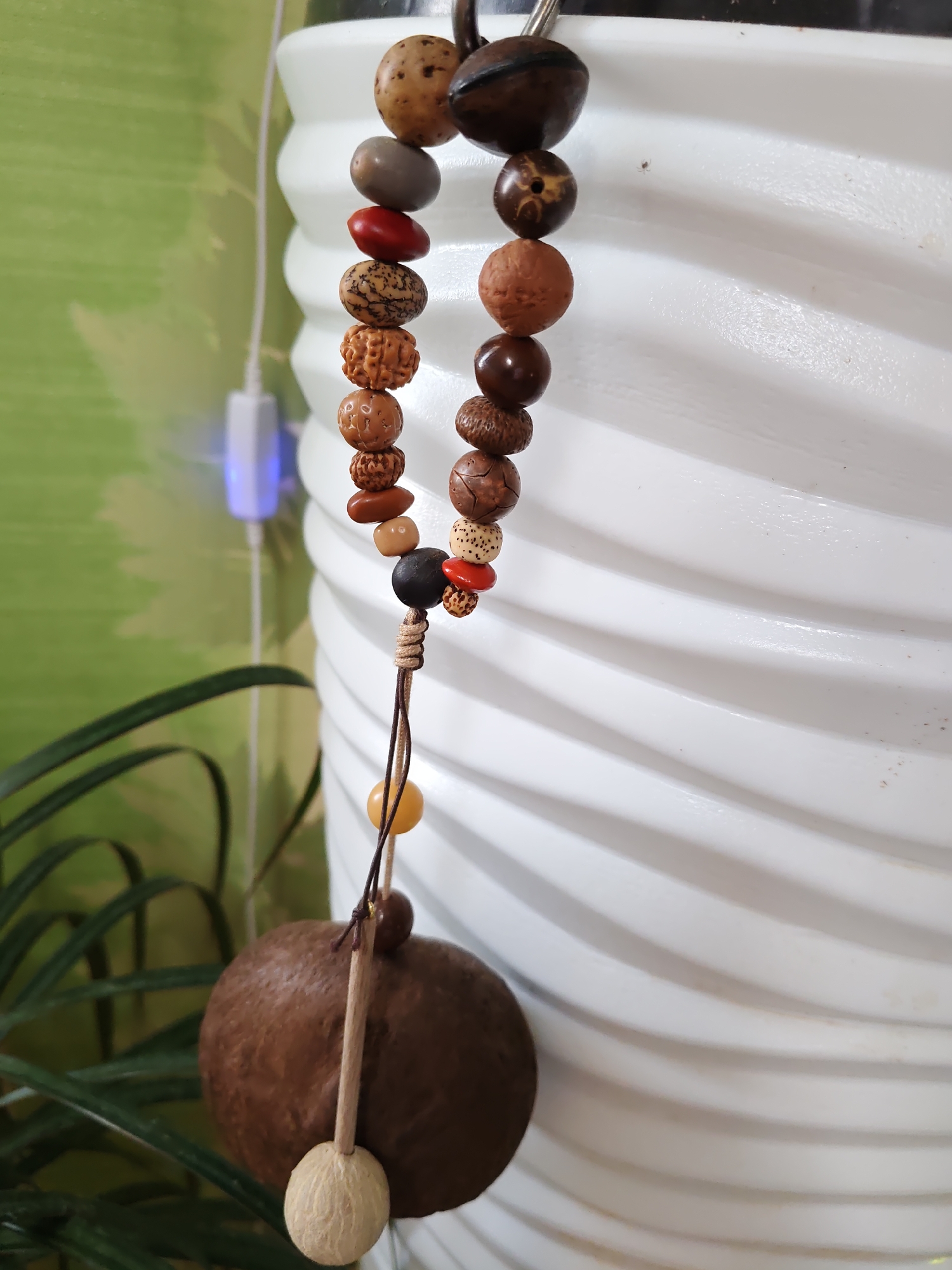 A close-up, eye-level, medium shot captures a string of wooden and natural beads hanging in front of a white, textured, cylindrical object. The beads, varying in size, shape, and color, are strung together with a brown cord. The top of the string is attached to a silver clasp. Below the beads, a large, round, brown object made of a natural material, possibly a coconut shell, hangs. A lighter colored, wooden piece is attached to the bottom of the shell. To the left of the hanging object, a green leafy plant is visible. In the background, a green wall with a subtle pattern is present, and a white electronic device with blue light is plugged into a socket on the wall. The scene appears to be indoors. The location is not specified in the image or its context to be Rostov-on-Don, Russia.