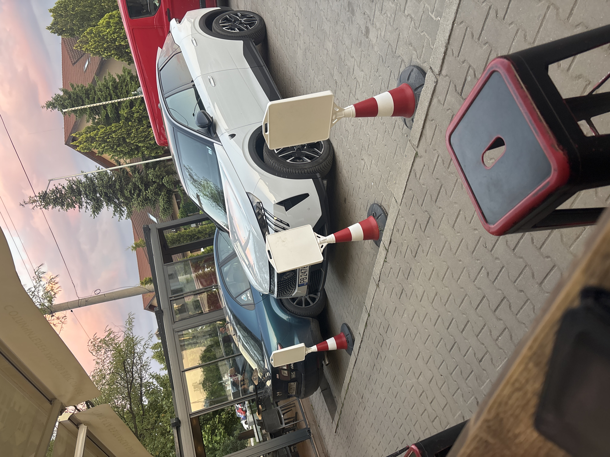 The image depicts two parked cars on a paved surface in Plovdiv, Bulgaria, separated by traffic cones with white placard holders. The first car is white, the second is blue. A red truck is partially visible behind the white car. A building with glass windows and a canopy is present. Part of a table and chair are visible in the foreground. The background includes trees and buildings under a sky with pinkish hues. No people are distinctly visible, though there may be reflections in the car windows. The location appears to be near a commercial establishment, possibly a cafe or restaurant. The license plates on the blue car are visible, showing "PB 2476" and "H 2287 AC".