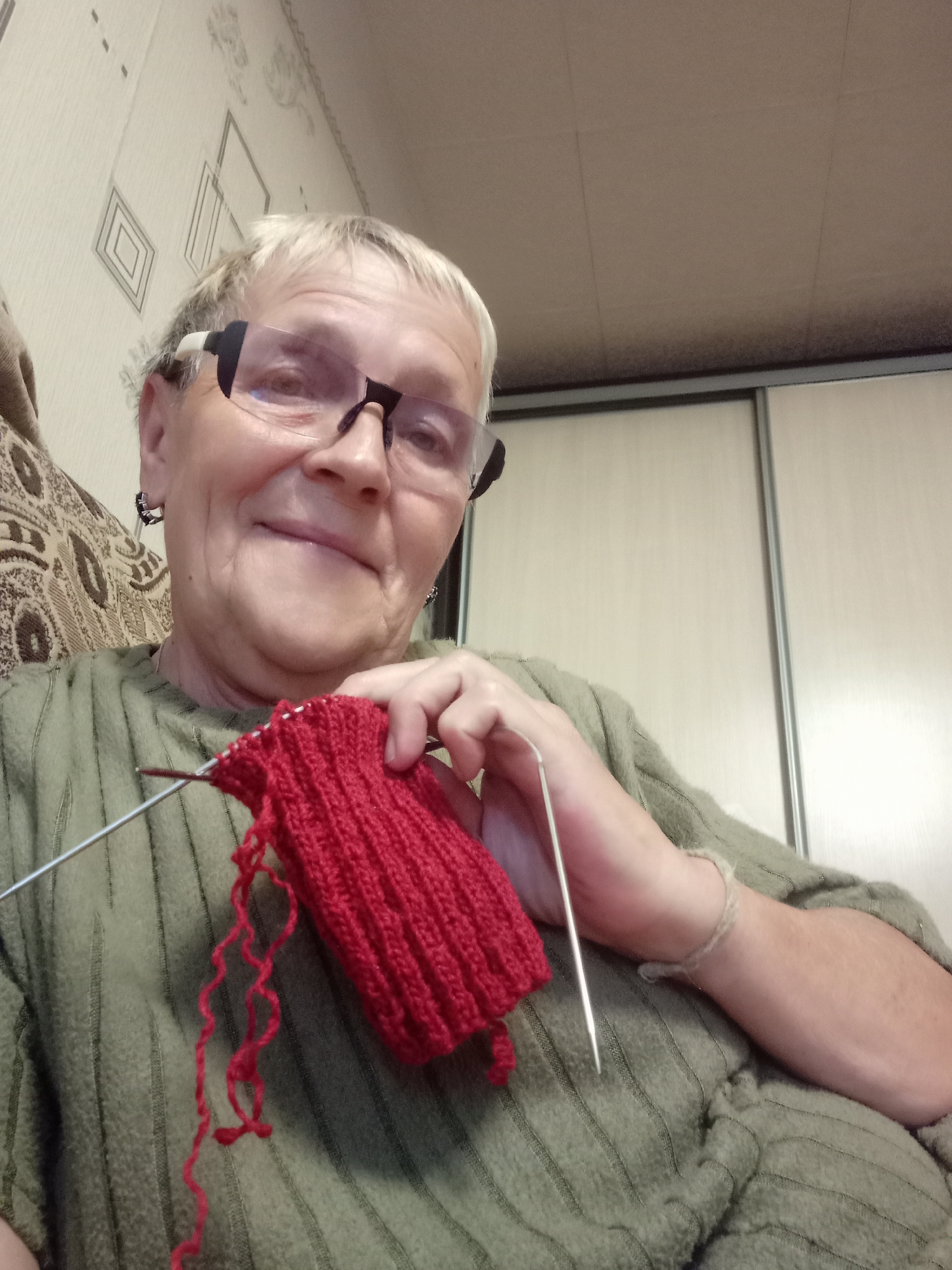 A woman with short blonde hair and glasses is knitting a red item on knitting needles. She is wearing a green knitted sweater. The background appears to be an indoor setting with a patterned wallpaper and a closet. The image is a selfie taken from a low angle, showing the woman's face and her knitting activity.