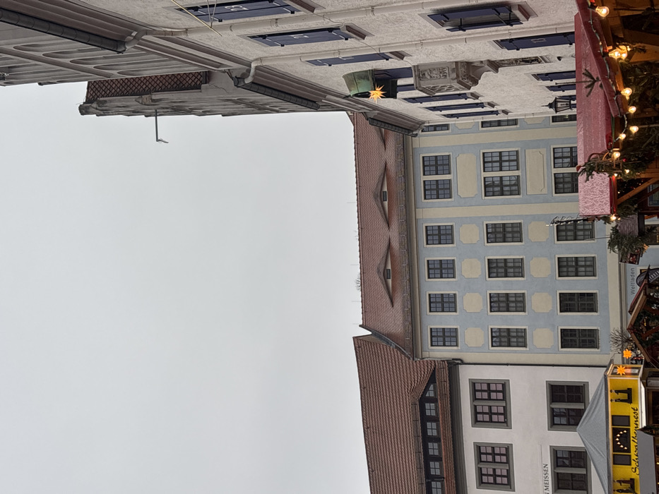 The image displays a daytime scene in the Markt (Market Square) of Meissen, Germany, under an overcast sky.
In the foreground right, a multi-story, light-colored building with a gabled roofline is visible. Its windows are fitted with dark blue shutters, three of which are numbered '21', '22', and '23', indicating an Advent calendar display. An ornate, carved stone balcony is positioned on the facade below one window. A gold, star-shaped light hangs from an archway or awning on this building. A metal downspout extends from the upper gabled section.
Adjacent to this, toward the left and center, are several other multi-story historical buildings. One building is light blue with cream-colored rectangular panels surrounding its dark-framed windows. Its reddish-brown tiled roof features two distinct oval "eyelid" dormer windows. To the far left, another off-white building with dark-framed windows also has a reddish-brown tiled roof with a dormer window. Partially visible text on its facade includes "MEISSEN" and "Schwalbennest."
The lower portion of the image shows several temporary market stalls. One on the far left has a yellow canopy with "Schwalbennest" text and string lights. Adjacent stalls feature dark canopies and are decorated with string lights and evergreen foliage. A sign with "Weltladen" is partially visible behind one stall. No individuals are discernible within the frame.