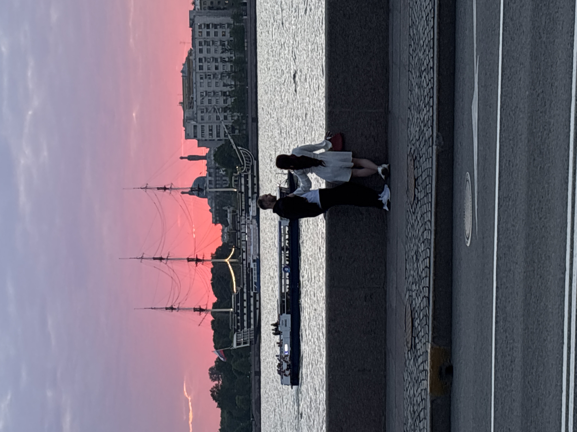 The image captures a picturesque urban waterside scene at sunset or twilight, characterized by a striking pink and purple gradient in the sky.
In the foreground, two individuals, a man and a woman, stand on a stone embankment overlooking a body of water. The man, wearing a dark top, light trousers, and white sneakers, is positioned slightly in front of the woman, who is dressed in a light-colored dress and shoes, and carries a red bag. They appear to be observing the view across the water.
The water itself, likely a river or large canal, shows ripples and reflections. On the opposite bank, a city skyline composed of various buildings is silhouetted against the vibrant sky. Dominating the mid-ground on the water is a large, multi-masted historic sailing ship, its rigging intricately silhouetted. A smaller, modern passenger boat, with several people visible on deck, is also present on the water, suggesting river tours or transport.
To the right of the two individuals, a cobblestone pathway runs alongside the embankment, further giving way to a paved road with clear white lane markings. The overall atmosphere is peaceful and scenic, enhanced by the dramatic sky. The lack of strong shadows and the rich sky colors strongly suggest evening, possibly during the "white nights" season. There is no visible text. Given the context, this is a view in Saint Petersburg, Russia.